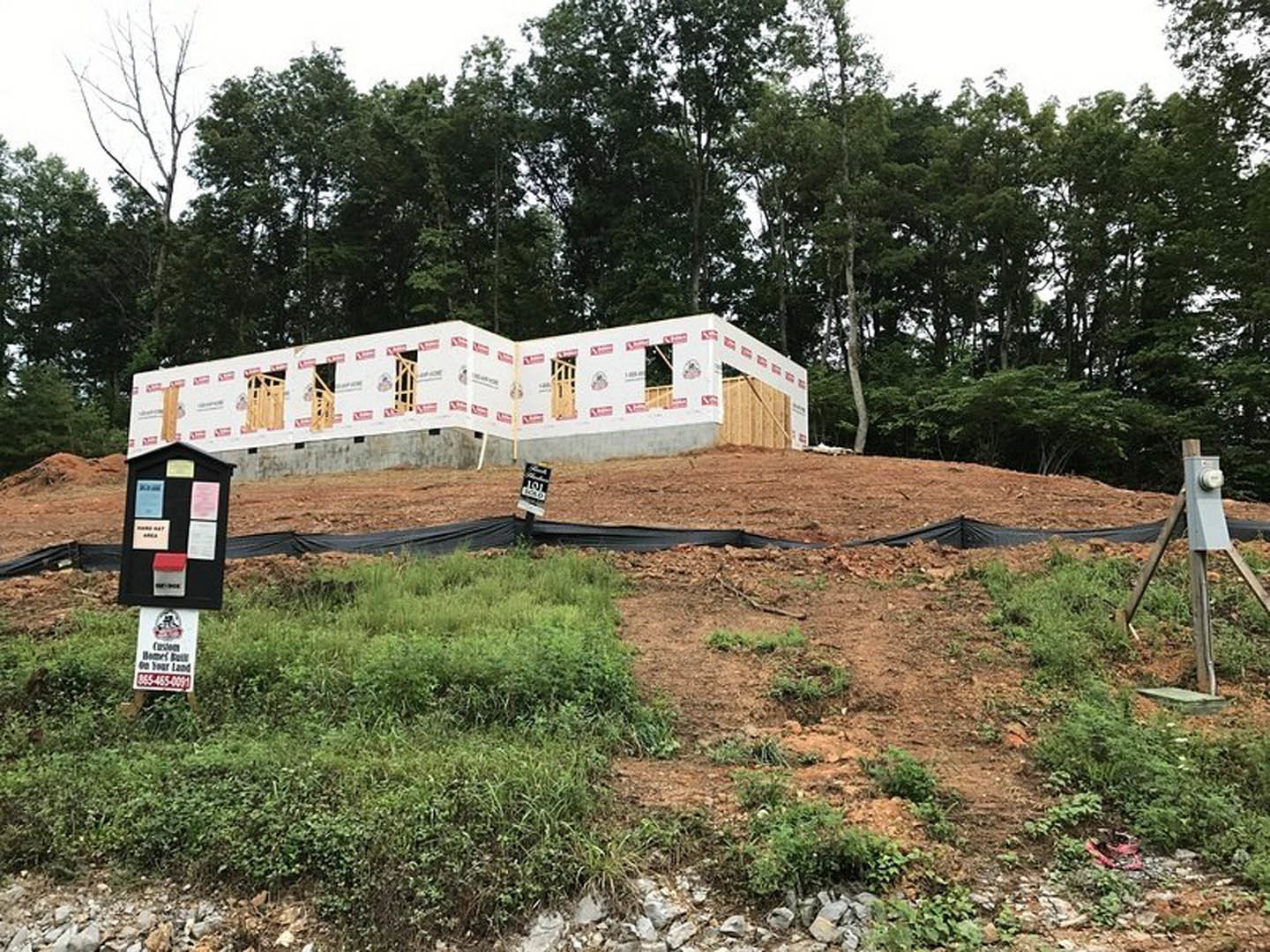 Partially built home framed with exposed wood, surrounded by dirt and grass, construction fencing, and trees in the background