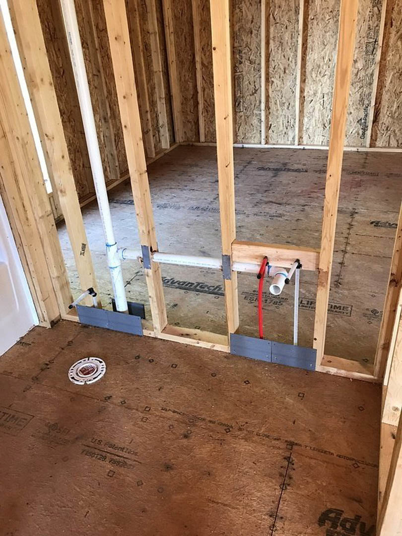 Unfinished room with exposed wood beams, plywood walls, visible plumbing pipes, and a hole in the subfloor