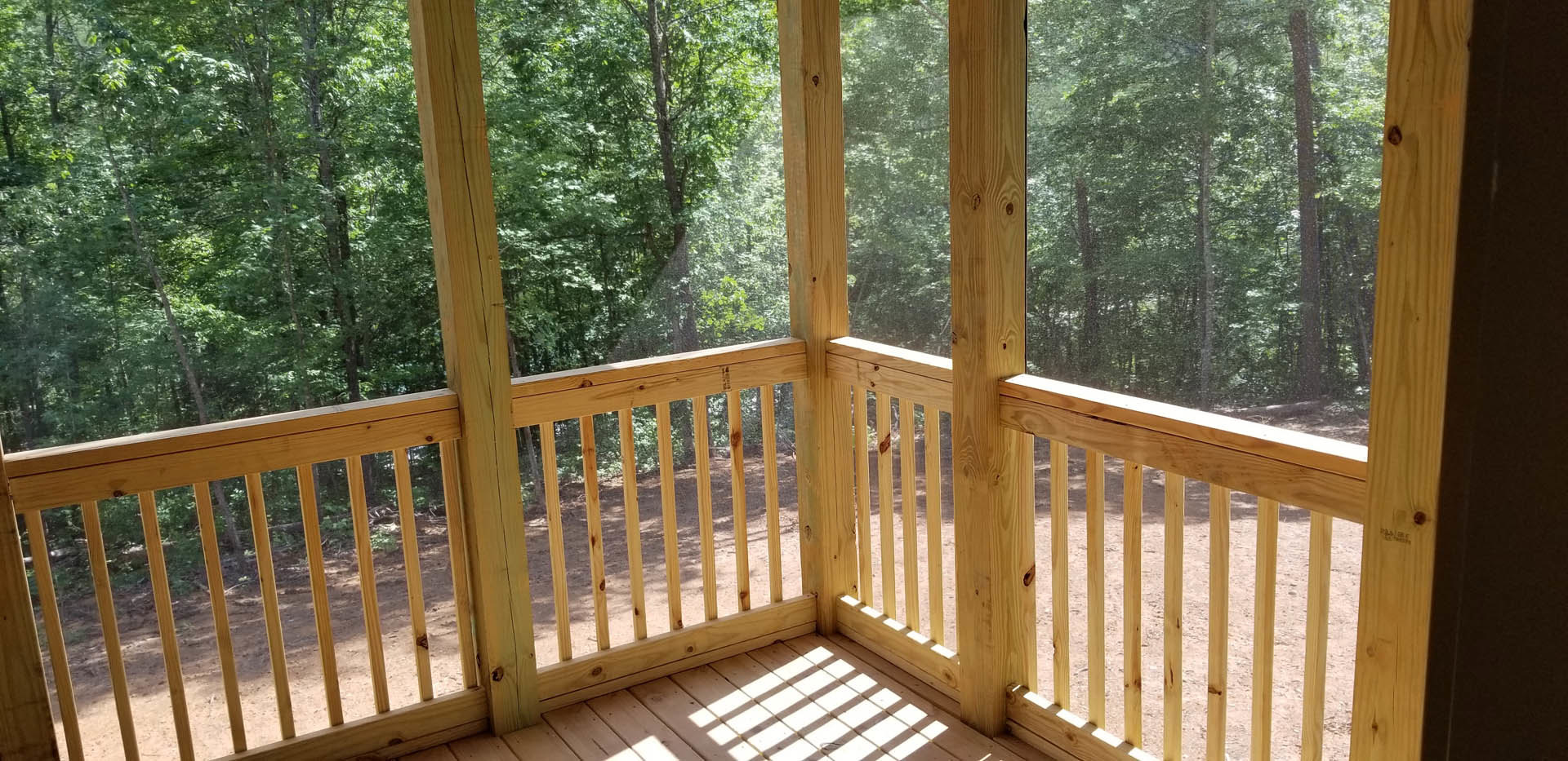 Wooden deck with railing overlooking leafy trees, sunlight filtering onto wood planks, exterior porch area with vertical posts and windows.