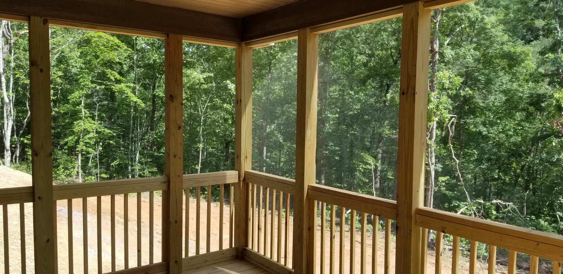 Wooden porch railing overlooking leafy trees, large window framing outdoor greenery, corner of room visible, natural wood finishes throughout