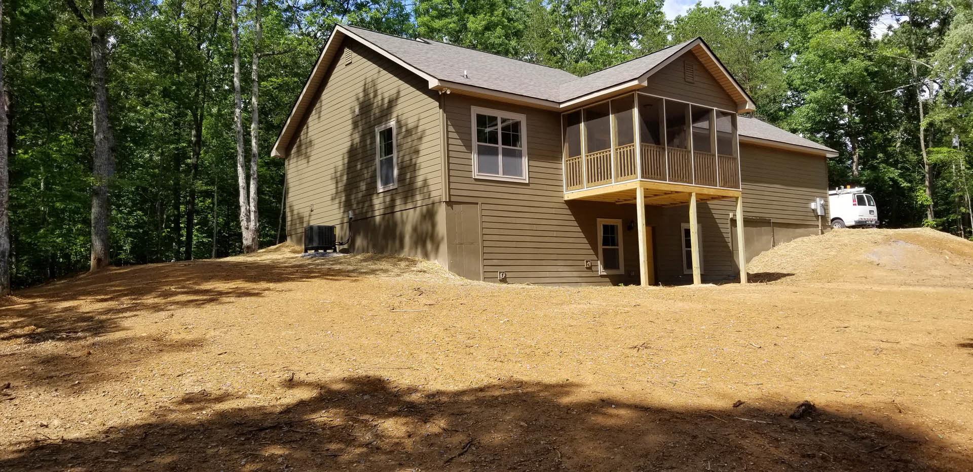 Two-story house with covered porch, white trim windows, dirt yard, and mature trees; white van parked nearby.