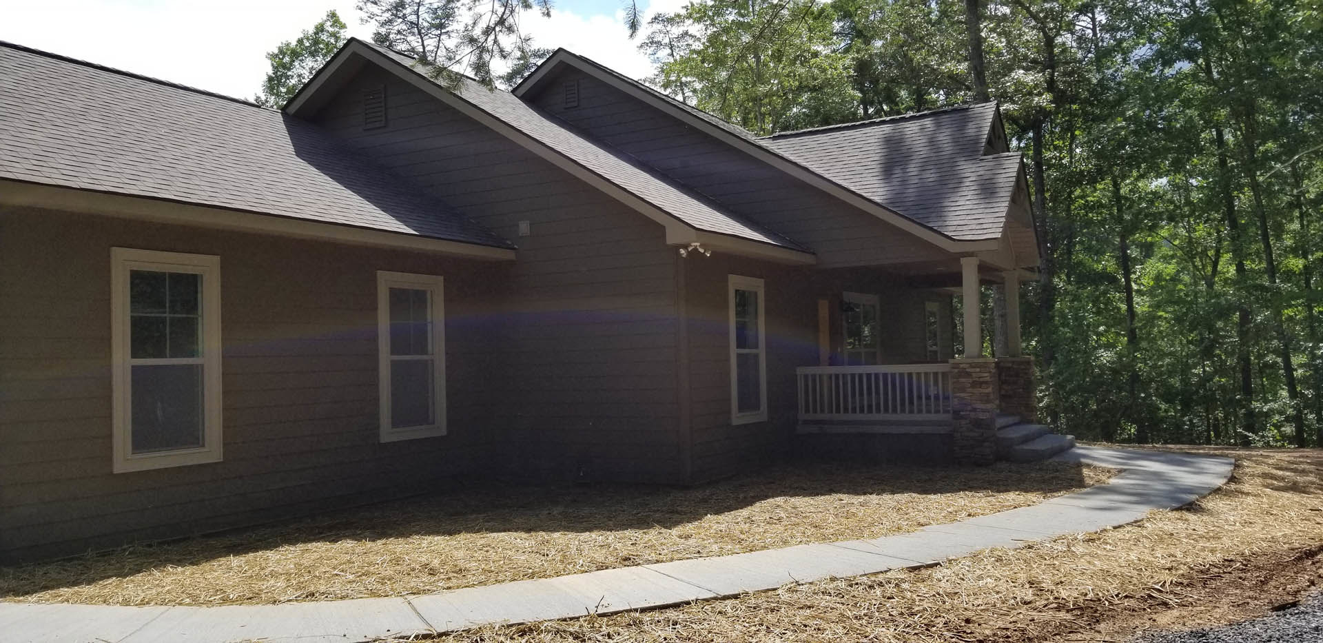 Two-story house with gray siding, white trim, covered front porch with white railings, large windows, concrete driveway, mature trees in background