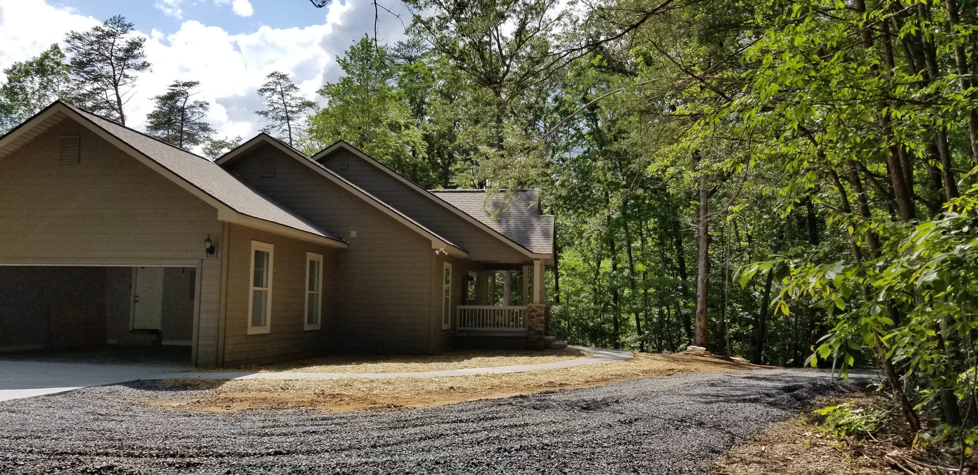 Two-story house with white-framed windows, attached garage, gravel driveway, mature trees with dense branches, and cloudy sky overhead