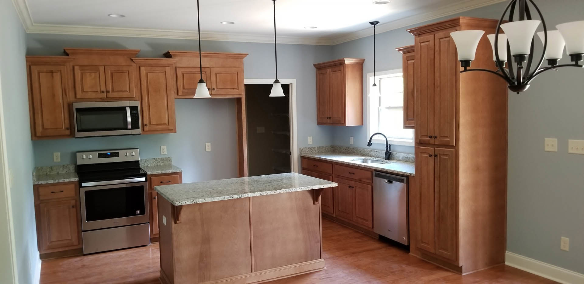 Kitchen with light wood cabinets, marble countertop, stainless steel oven with black door, built-in microwave, modern chandelier, and sink; partial view of doorway in background.