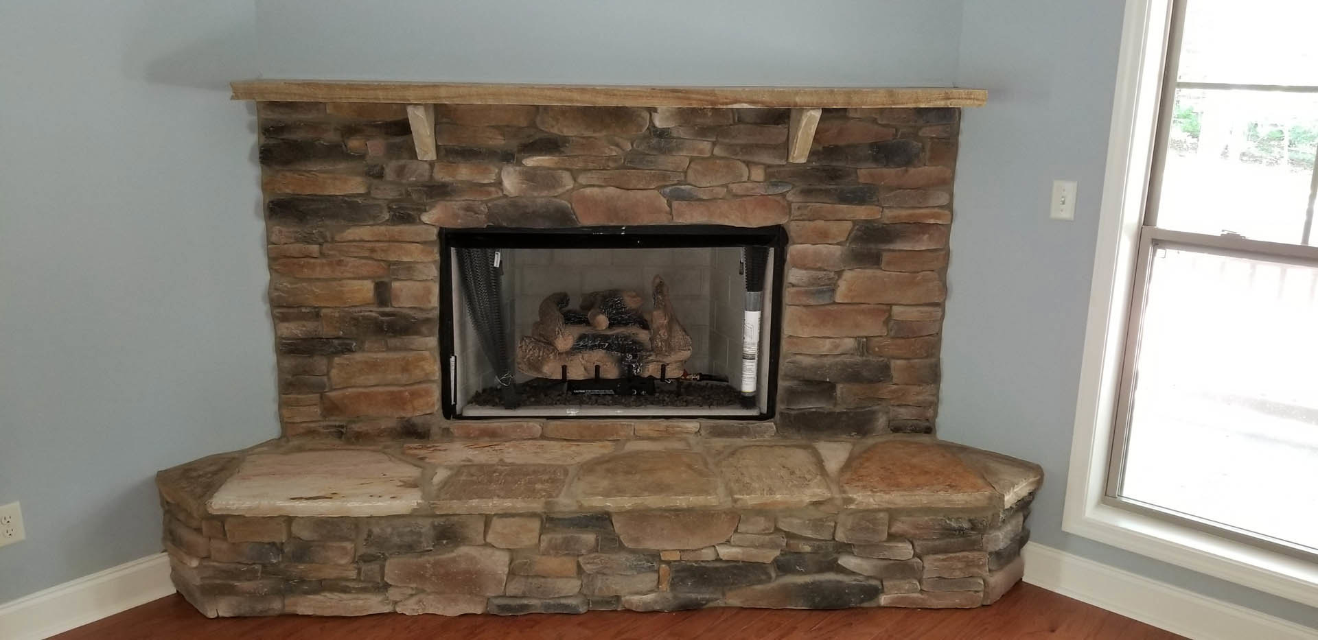 Stone fireplace with wood mantel shelf, stacked logs in hearth, fire screen, white-framed window, and light switch on adjacent wall
