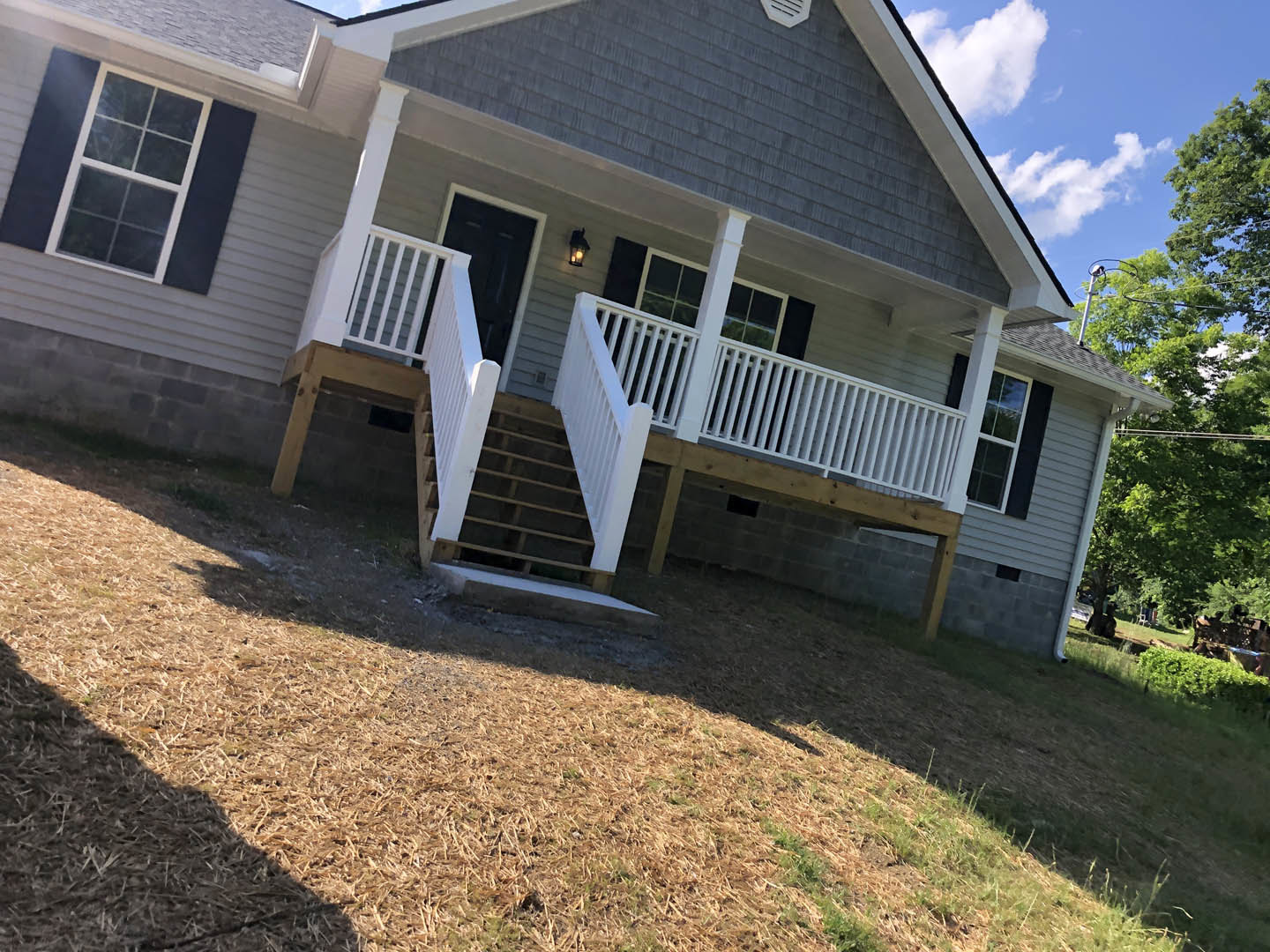 White siding house with covered porch, white staircase and railing, black front door, white-framed window, surrounded by trees under blue sky