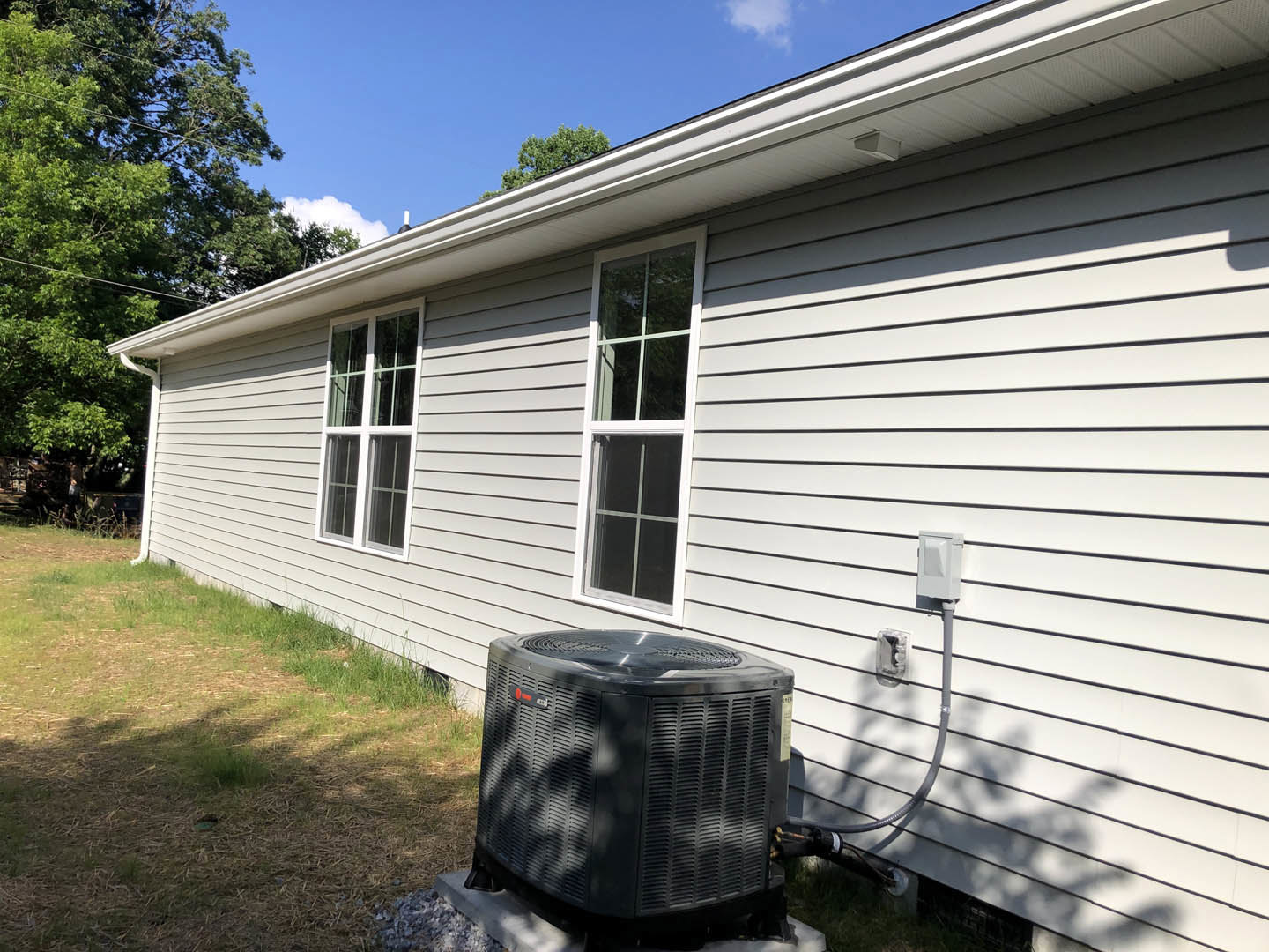 Grey siding home exterior with white-framed window, black heat pump unit positioned beside the house, green grass lawn, and trees in background