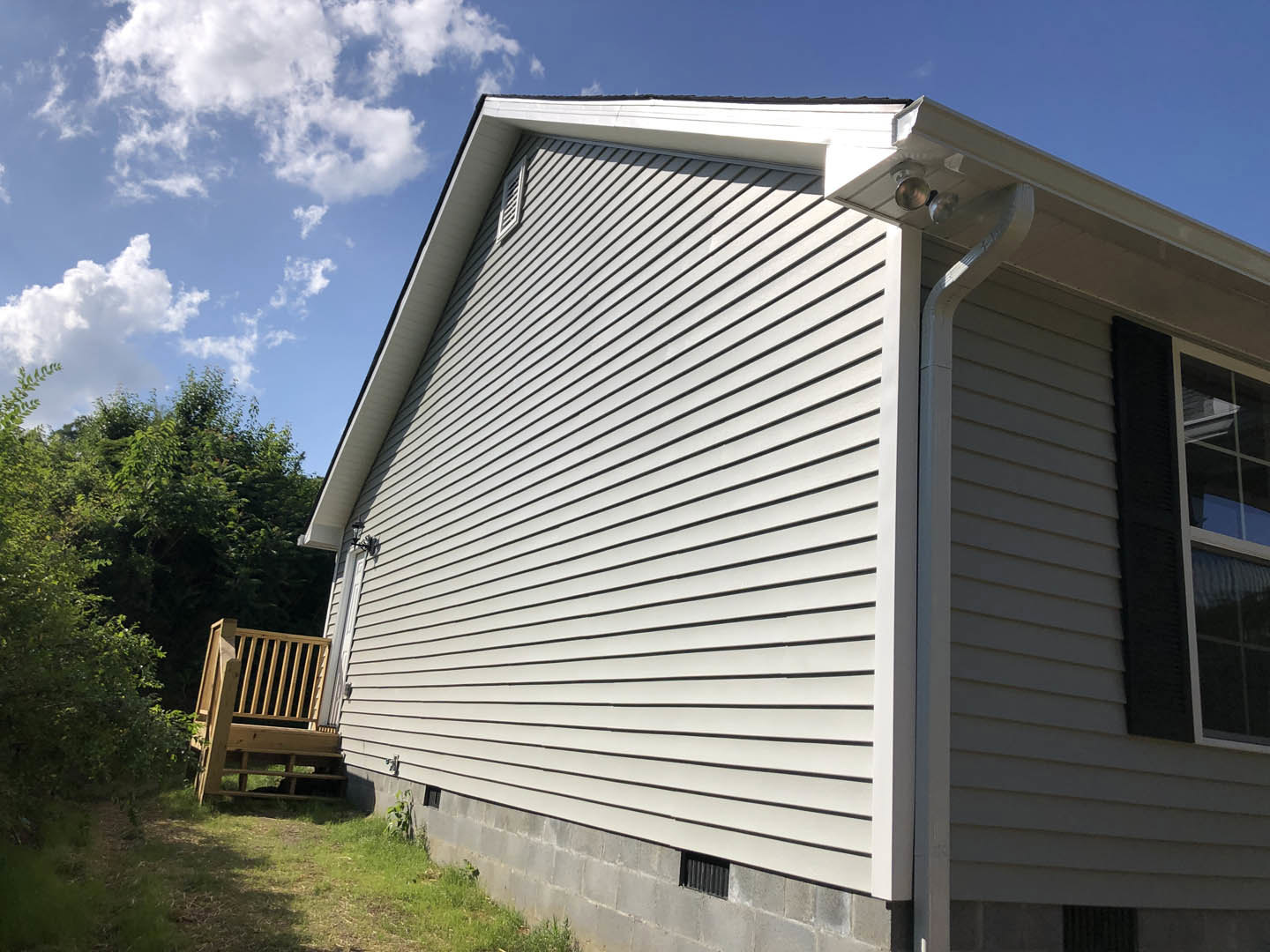 Two-story home with light siding, covered wooden porch with railing, elevated deck, white-trimmed windows, black roof tiles, and landscaped yard with trees under partly cloudy sky