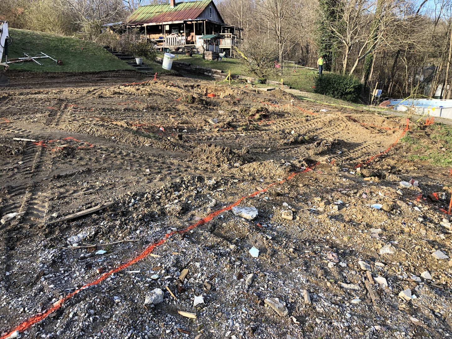 Single-story house with green roof surrounded by trees, set on a dirt field with patches of grass and soil under a clear sky