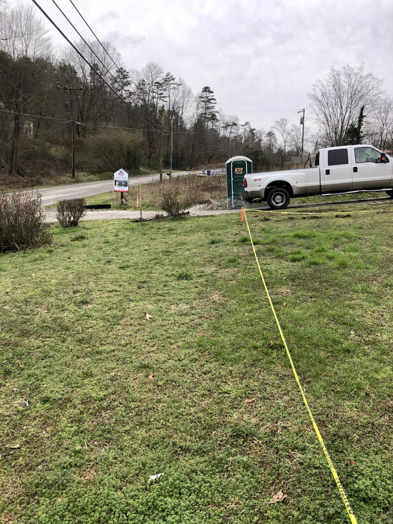 White truck parked on grassy lot with long yellow tape stretched across grass, portable toilet nearby, bush and trees in background.