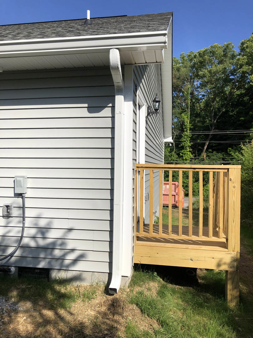 Wooden deck with railing attached to house, bordered by a fenced yard and gate, surrounded by green grass and mature trees under a sunny sky; visible siding, gutter, and outdoor