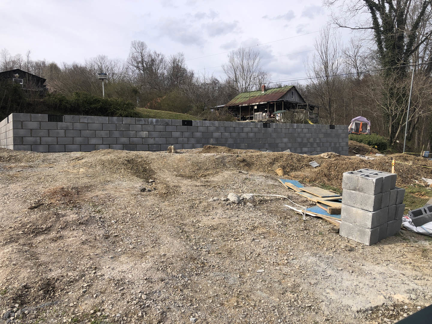 Grey concrete block wall with rectangular opening, stack of bricks on dirt field, green-roofed house and white tent in grassy background, scattered tools and plants.