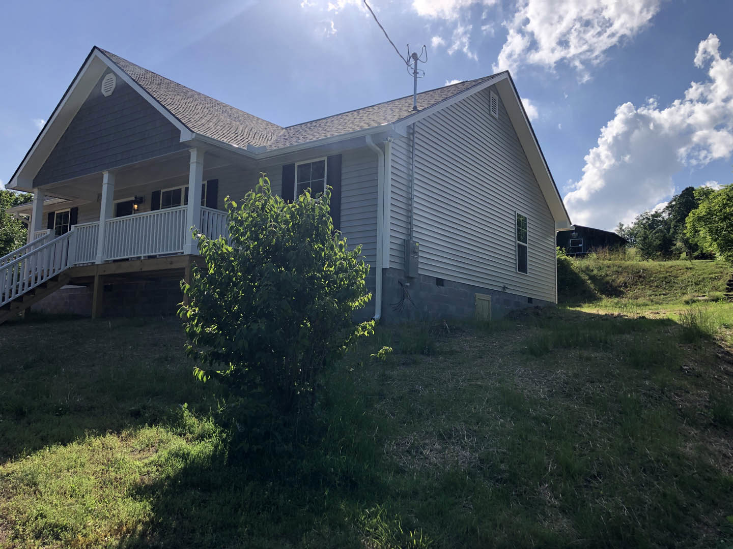 Two-story home with white-framed windows and white staircase railing, situated on a grassy hill with a large tree in the backyard under a partly cloudy sky