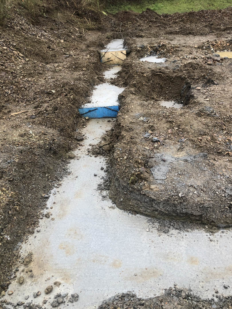 Dirt trench with multiple holes and patches of soil, surrounded by grass and plants, prepared for residential foundation footers