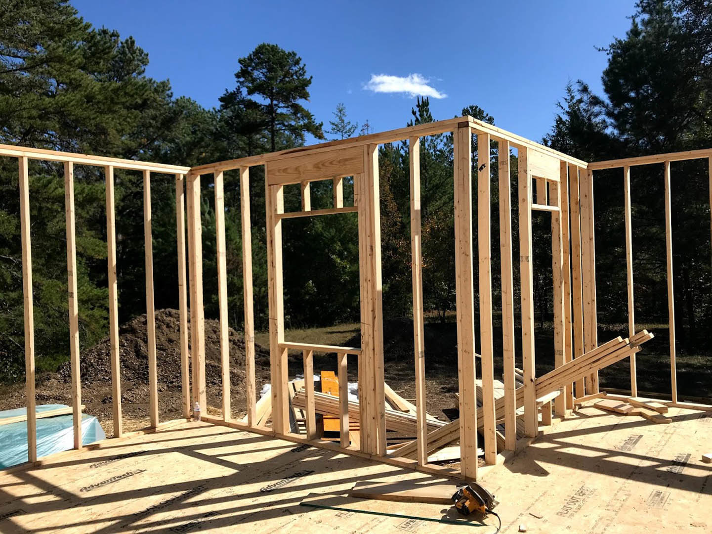 Wooden house frame under construction with blue tarp, piles of dirt and lumber, fenced yard, trees, and partly cloudy blue sky