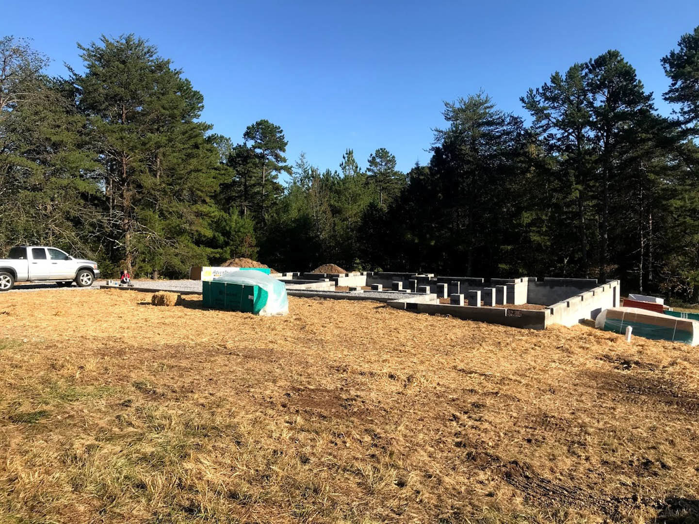 Grassy building site bordered by tall trees, white truck parked on dirt road, green tent and container with white cover visible in open field