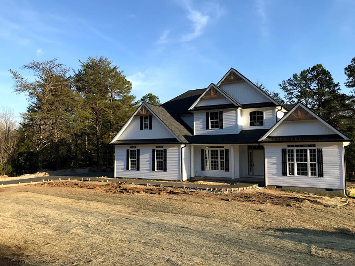 Two-story house with black roof, white window frames, surrounded by trees, set beside a dirt road under cloudy sky