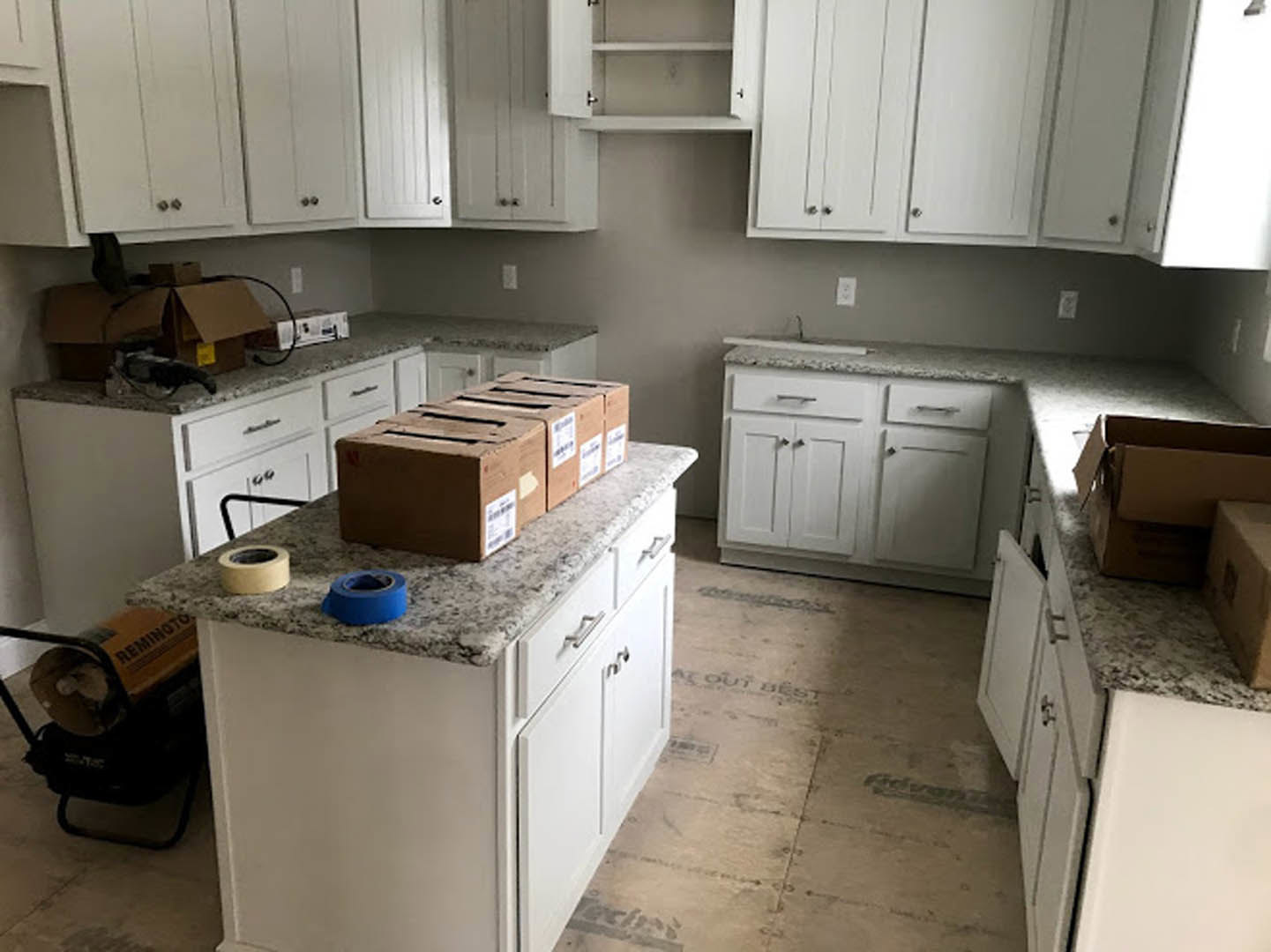 White kitchen cabinets with cardboard boxes and a roll of blue tape on the countertop, hardwood flooring, stainless steel appliances, and a cart with rolled cardboard