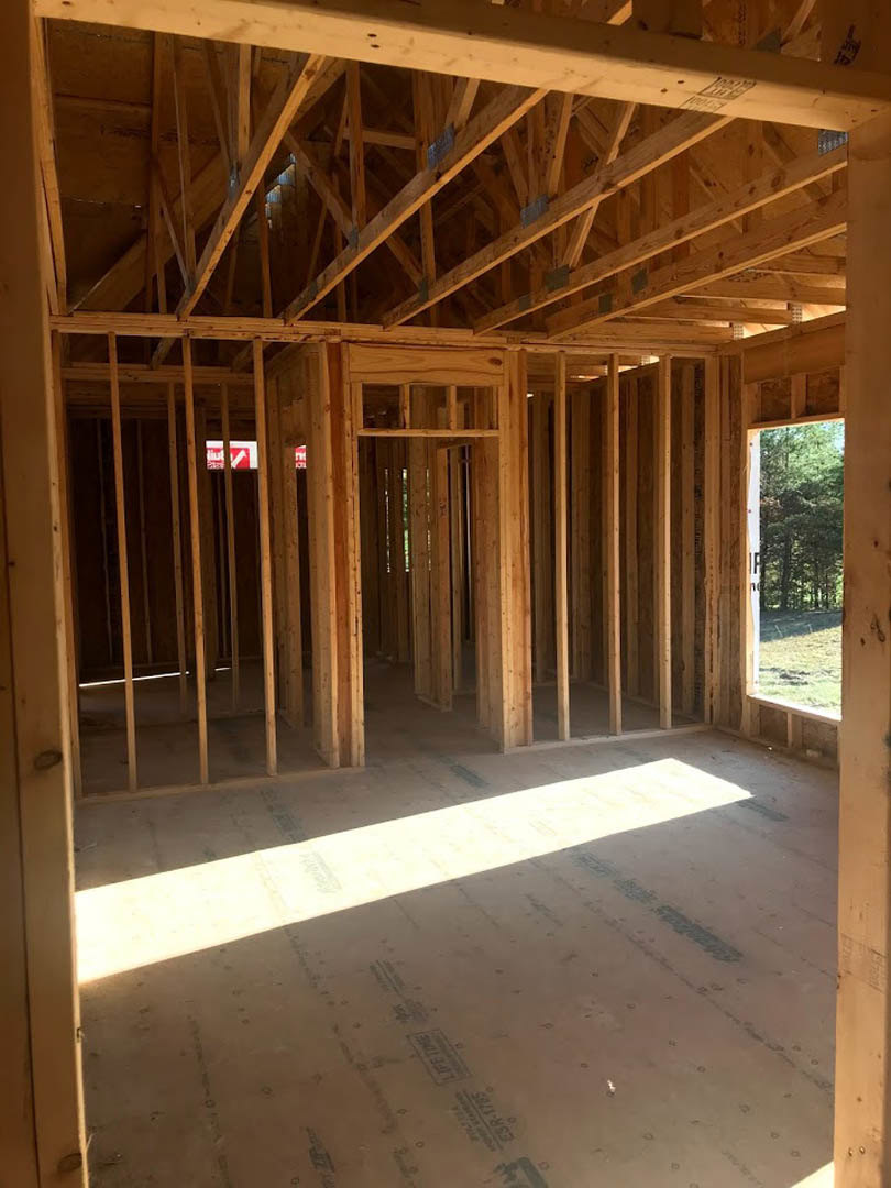 Exposed wood framing and roof trusses inside a house under construction, unfinished floor with lumber beams, sunlight streaming through open window, forest visible outside