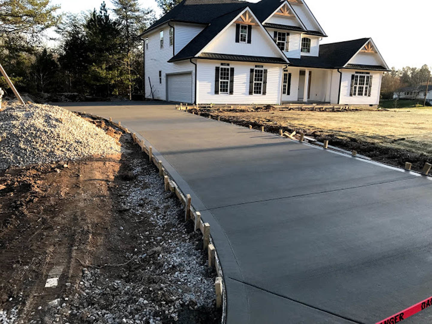 Concrete driveway leading to a modern house with a triangular roof, multi-pane windows, white gravel landscaping, and mature trees in the background