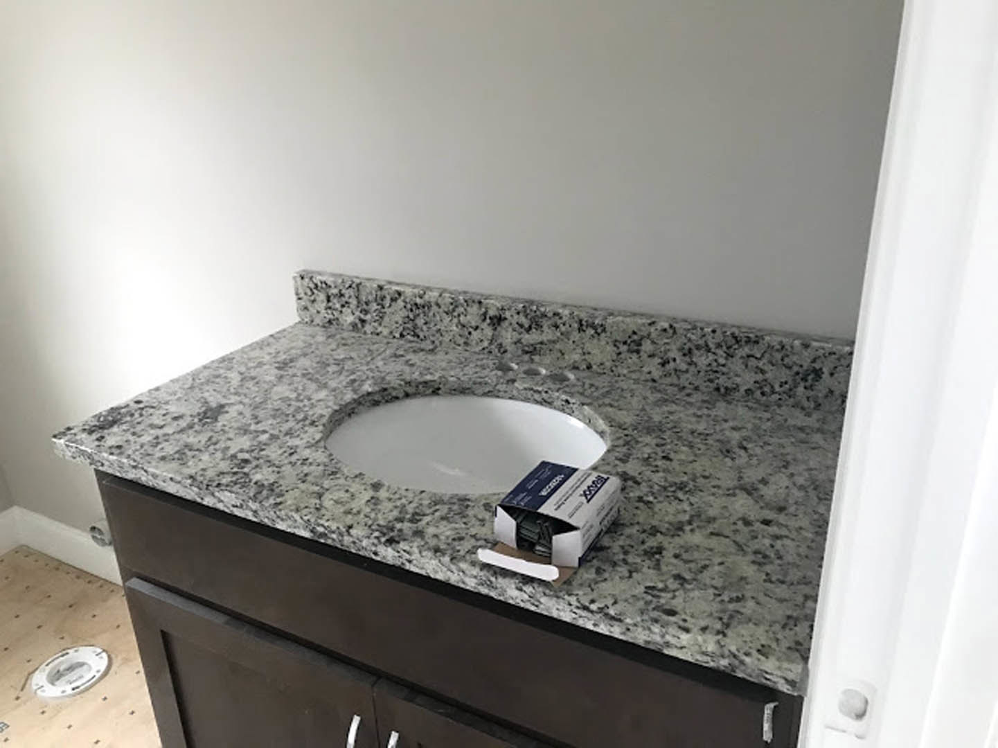 Undermount sink set in a marble countertop beside a white toilet, with a box of paper and a round wall clock displaying black numbers, surrounded by white cabinetry and tiled