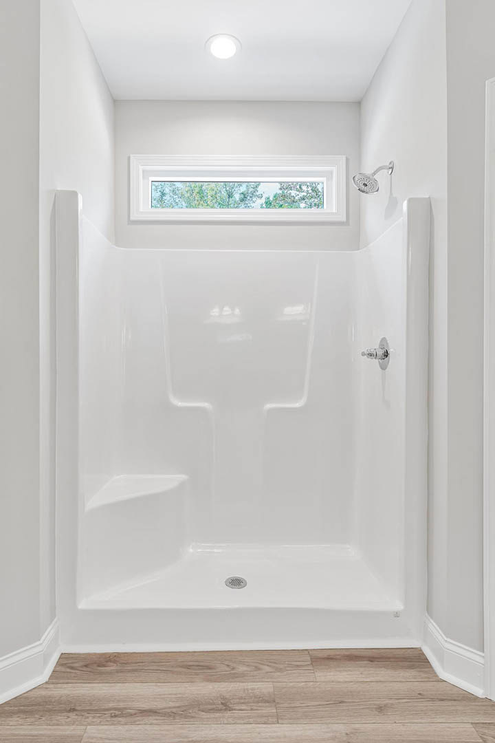 White bathroom shower with glass enclosure, window overlooking trees, wood floor with white trim, chrome shower head and nozzle.