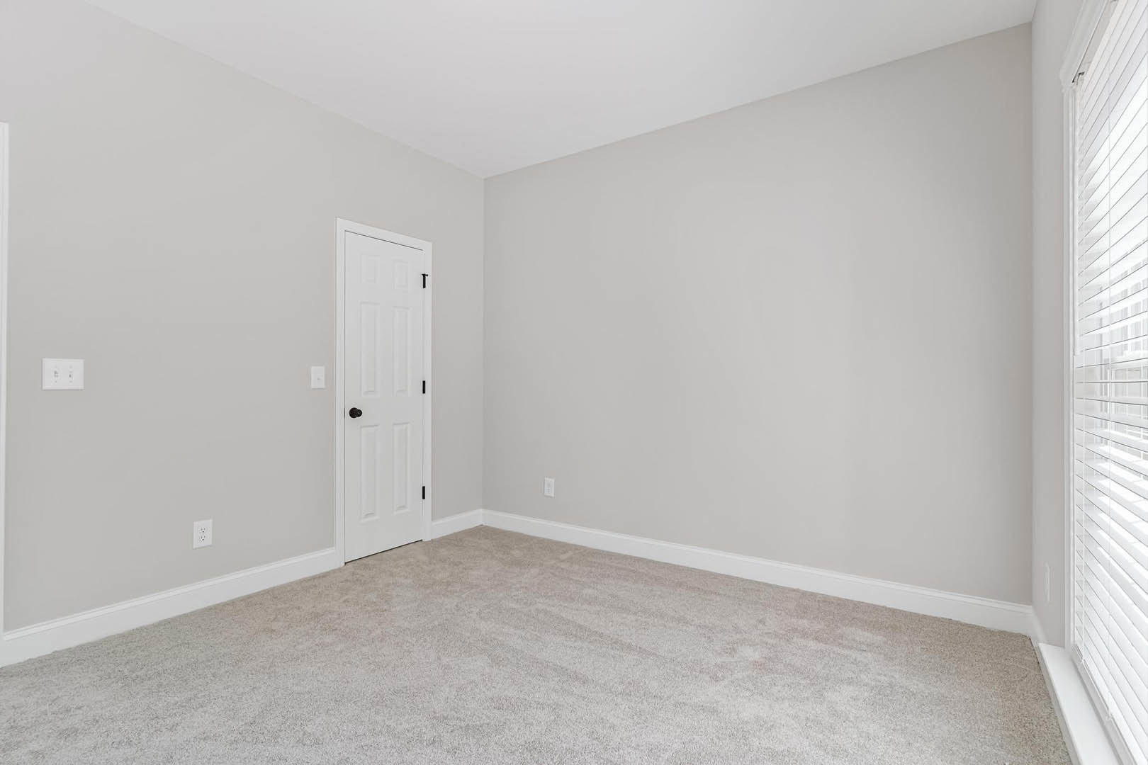 White carpeted room with white walls, white door featuring black knob, wall outlet with black buttons, and window with white trim.