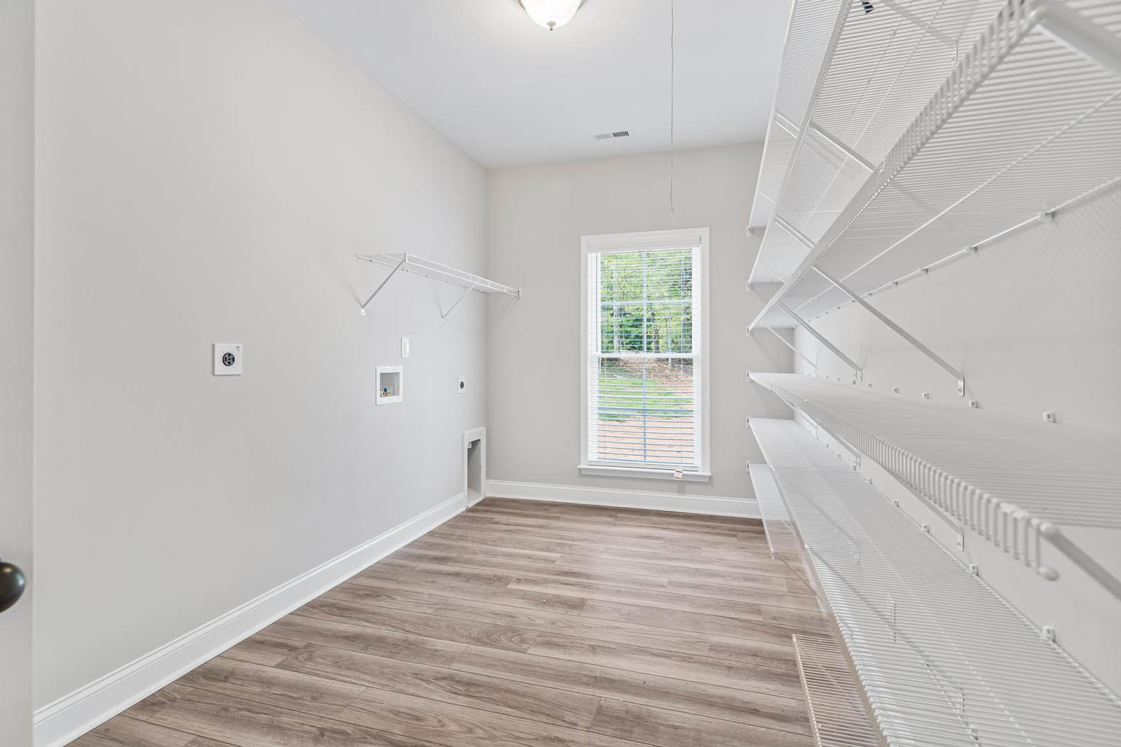 Wood flooring and white walls in an empty room with built-in white shelves featuring metal rods, a window with blinds, and a white electrical outlet.