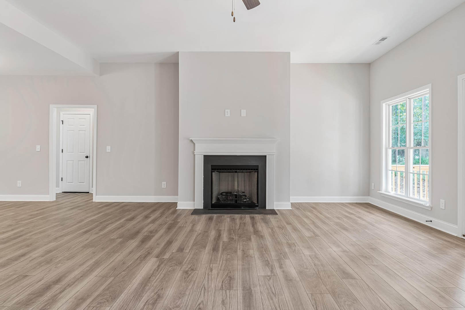 Living room with white-trimmed fireplace, wide plank hardwood floors, white door with black hardware, and large multi-pane windows.