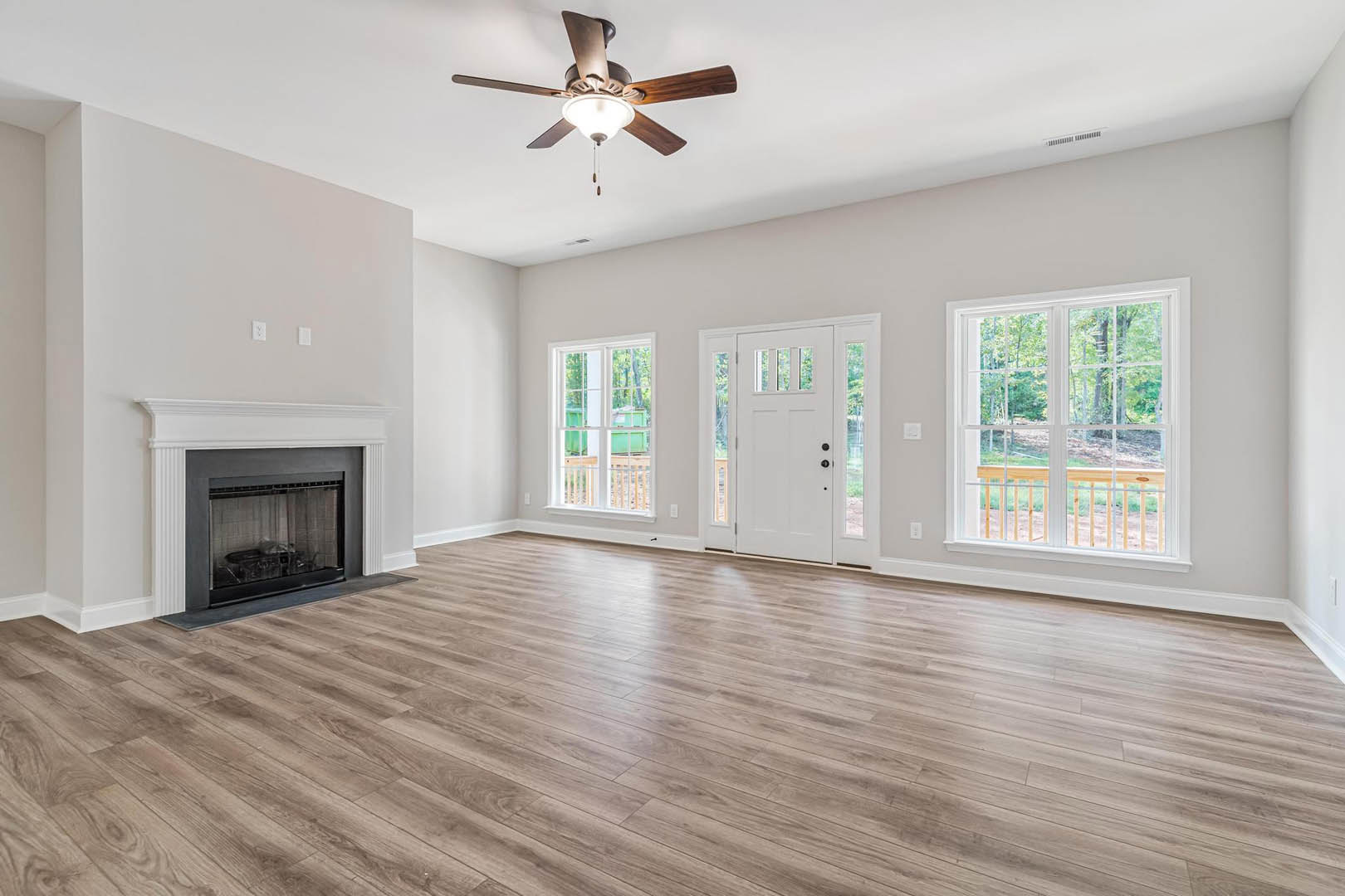 Living room with hardwood flooring, ceiling fan with light, glass door fireplace, large window overlooking trees, and white door with glass panels