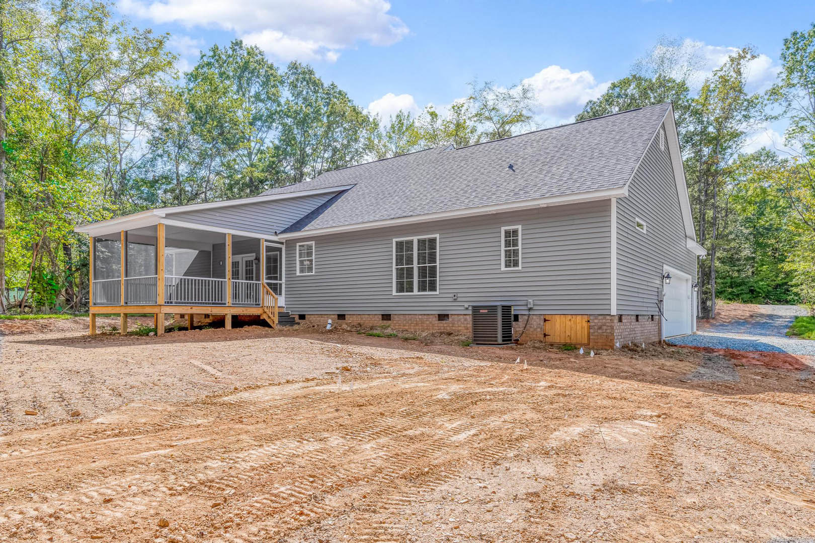 White cottage-style home with covered porch, wooden deck, dirt driveway with tire tracks, metal utility box with red sign, wooden fence, surrounding trees and cloudy sky