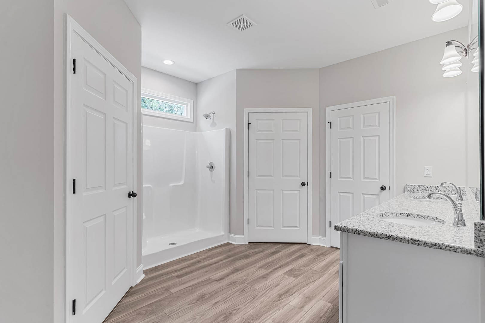 Bathroom with white paneled doors featuring black handles, wood flooring with a drain, white shower enclosure with silver hardware, and a white sink with chrome faucet.