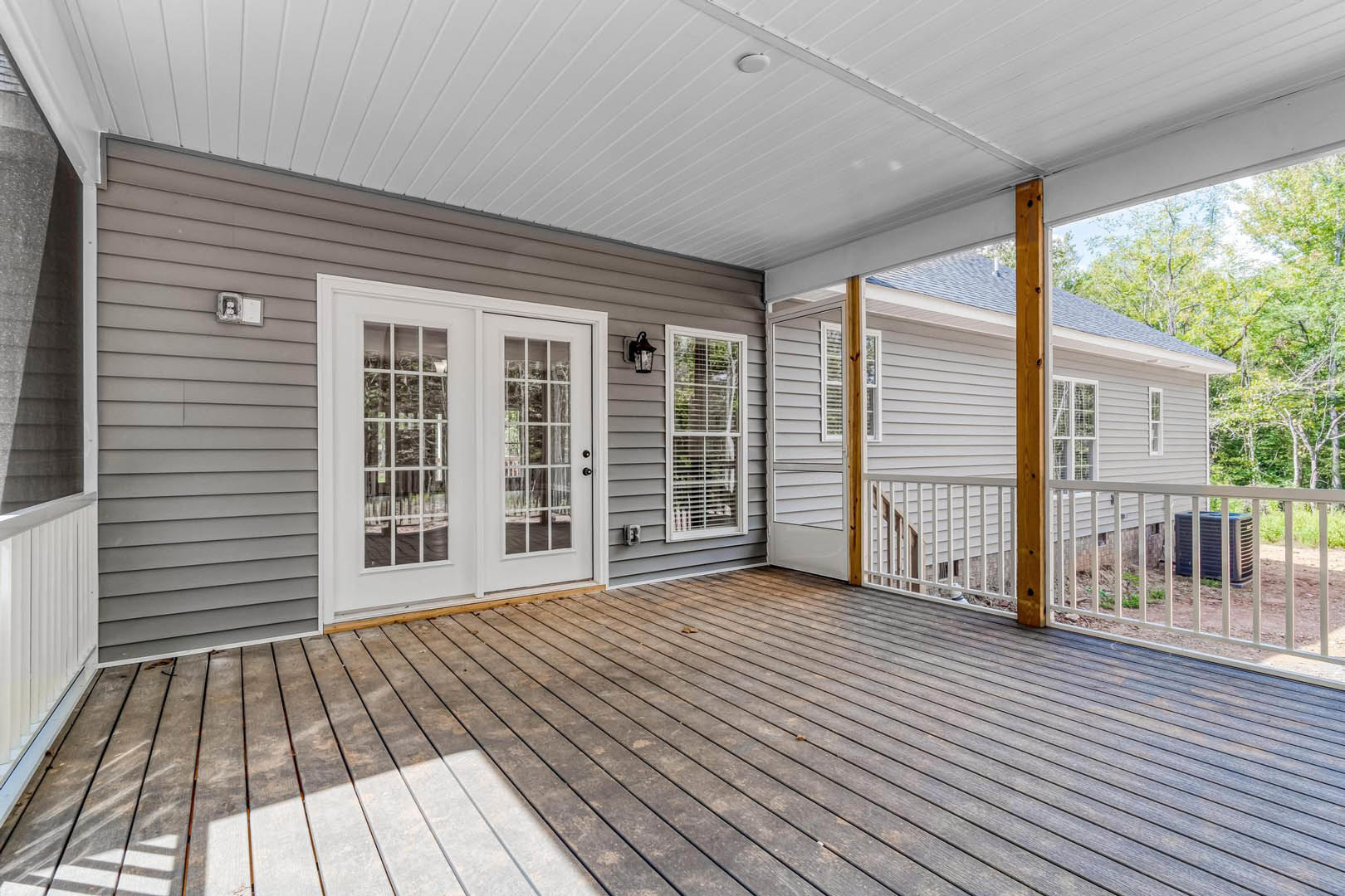 Wooden deck with white double doors featuring glass panes, covered porch ceiling, adjacent window, white fence enclosing black air conditioner unit, and exterior composite