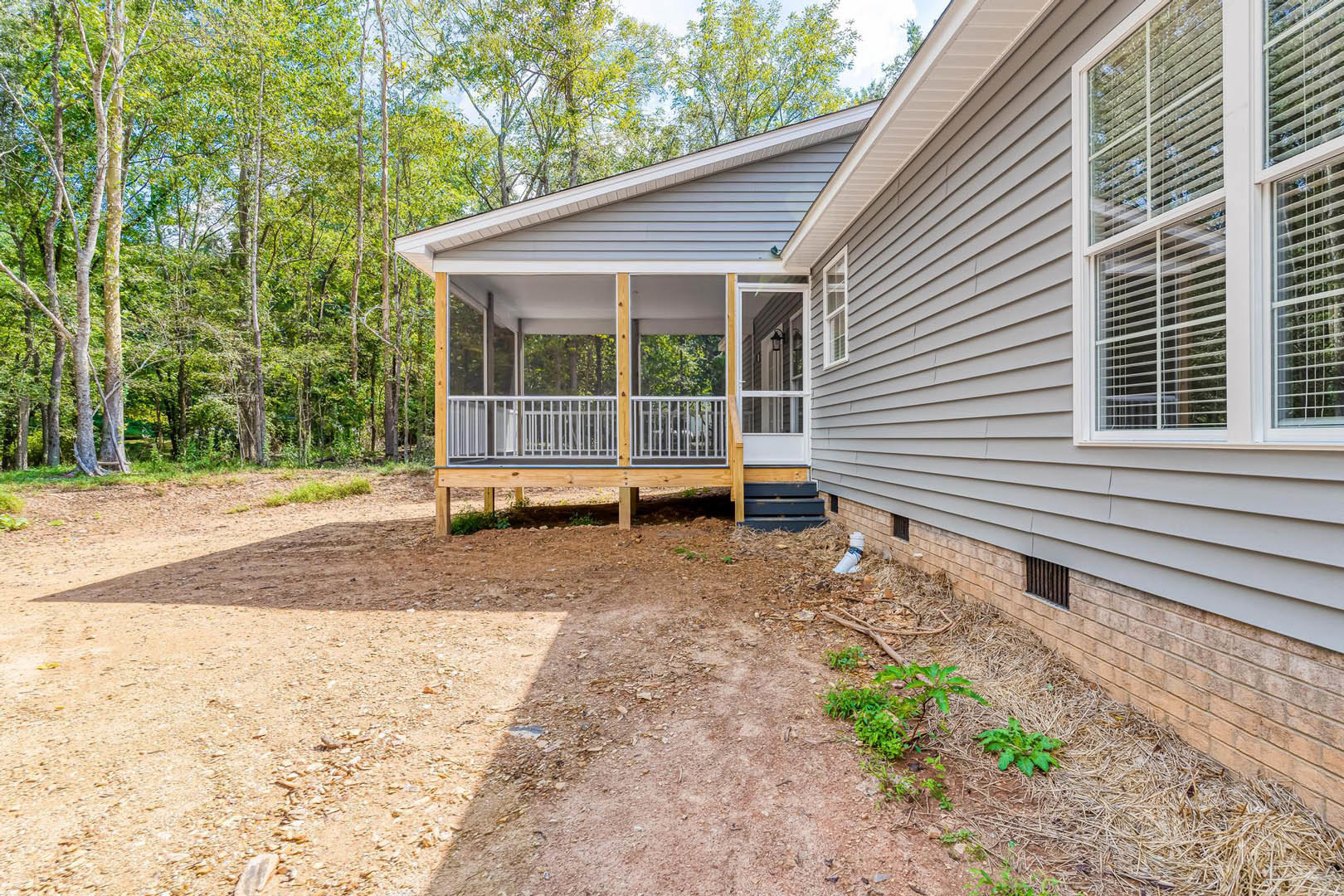 Two-story house with light siding, yellow front door, white porch railing, window with blinds, dirt path leading to entrance, surrounded by trees and yard.