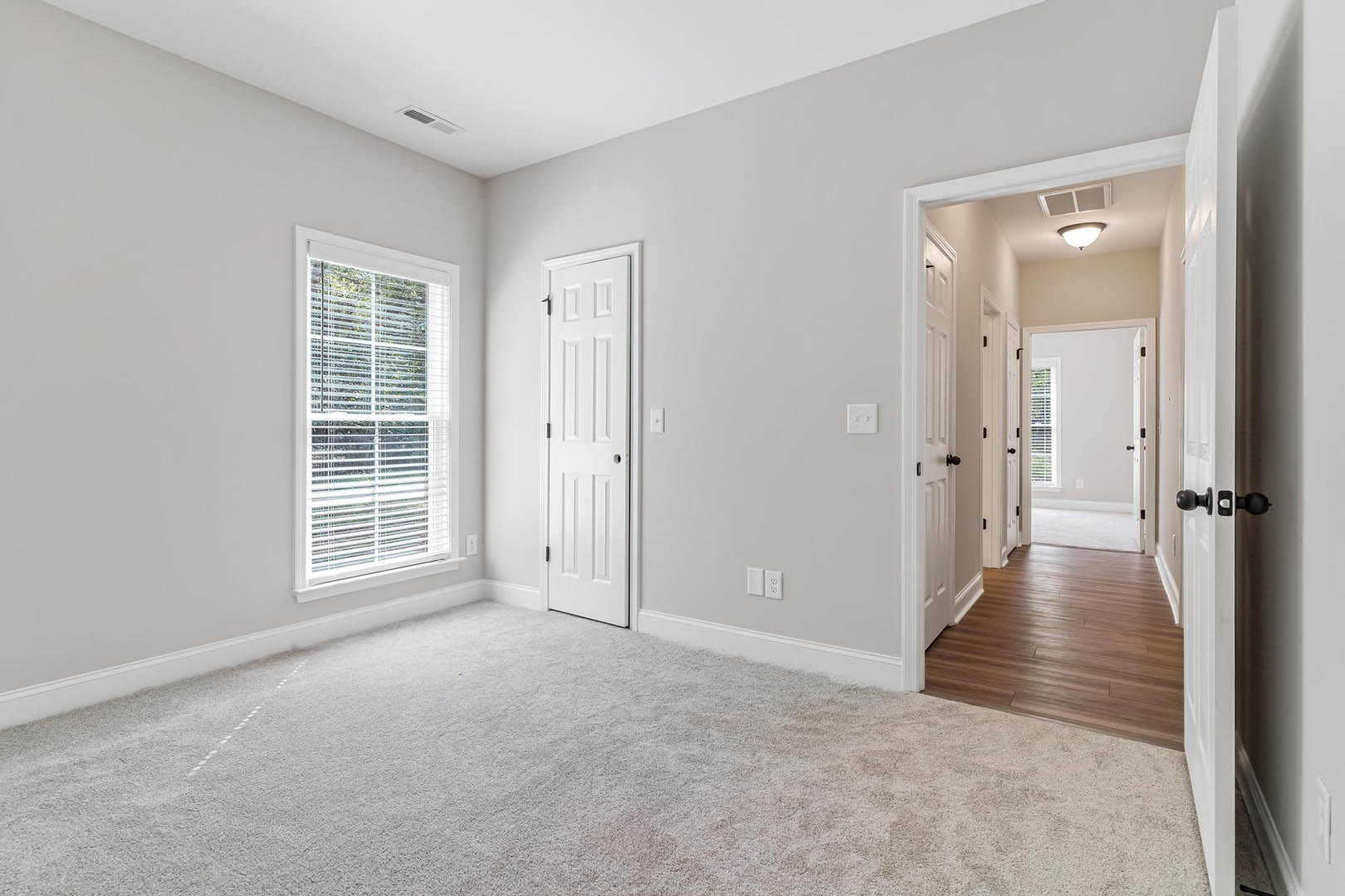 White-walled room with plush white carpet, window fitted with horizontal blinds, white door featuring a round knob, and wood laminate flooring visible near the entry.