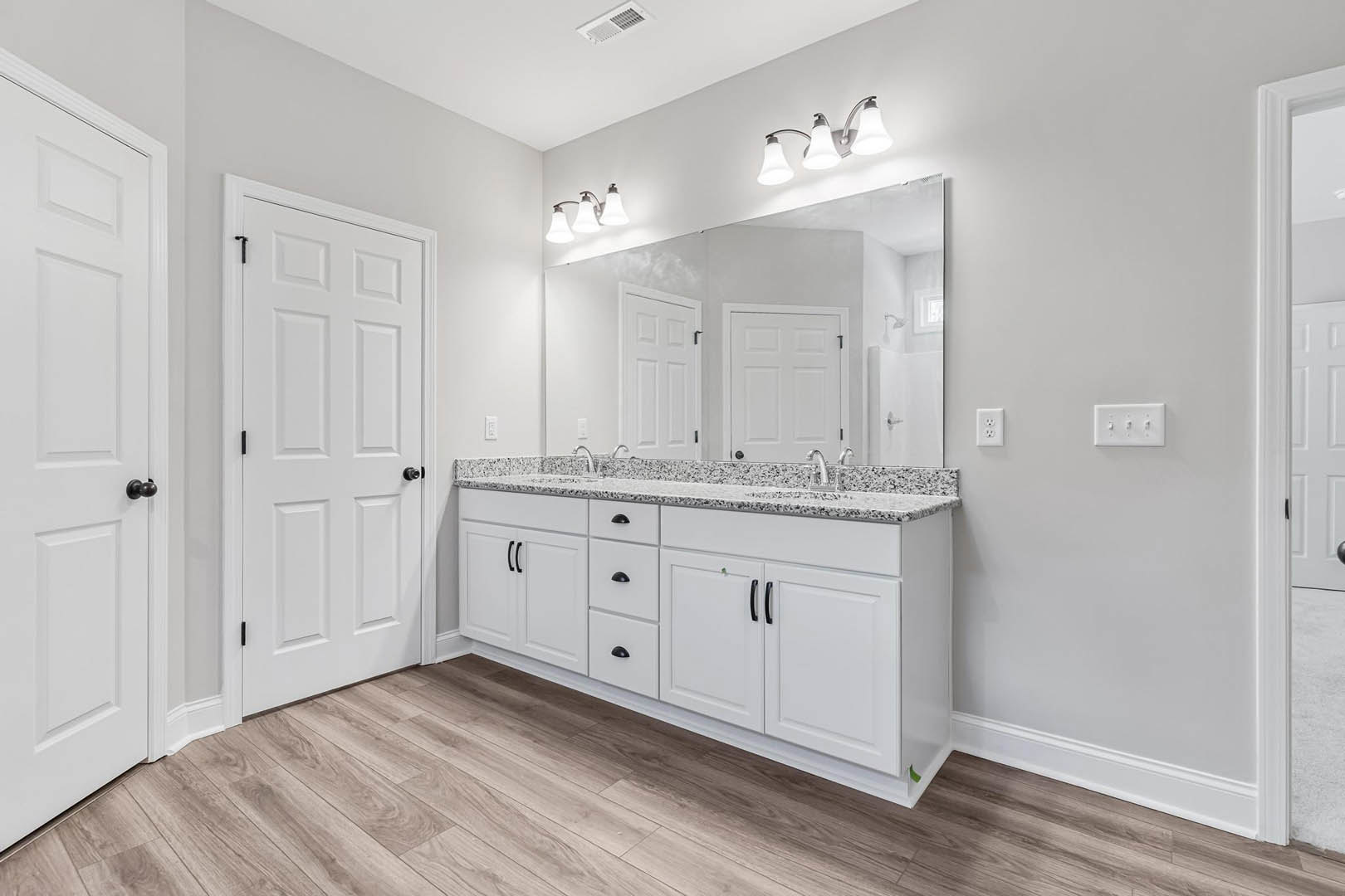 Bathroom with a large framed mirror above a white countertop, white door with black knobs, multi-switch white light switch, wall vent, and three-light fixture.