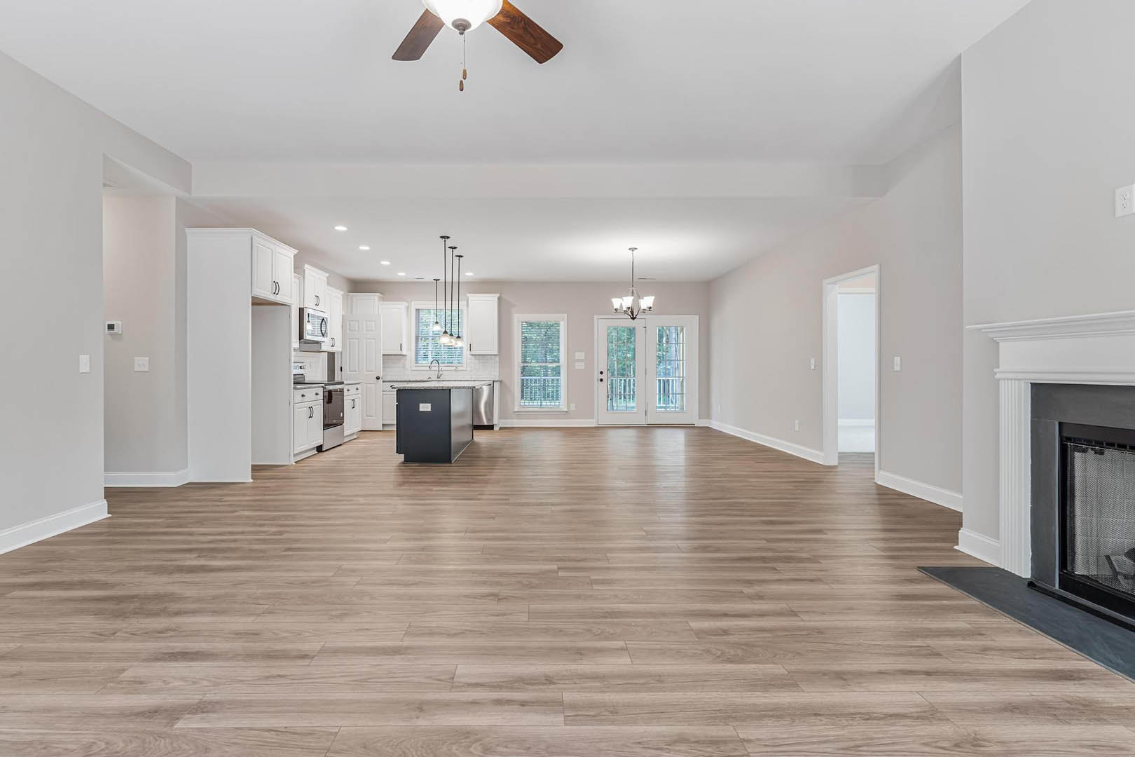 Spacious open-concept living area with hardwood flooring, white plaster walls, ceiling fan with light fixture, window with blinds, white double doors with glass panes, and a black