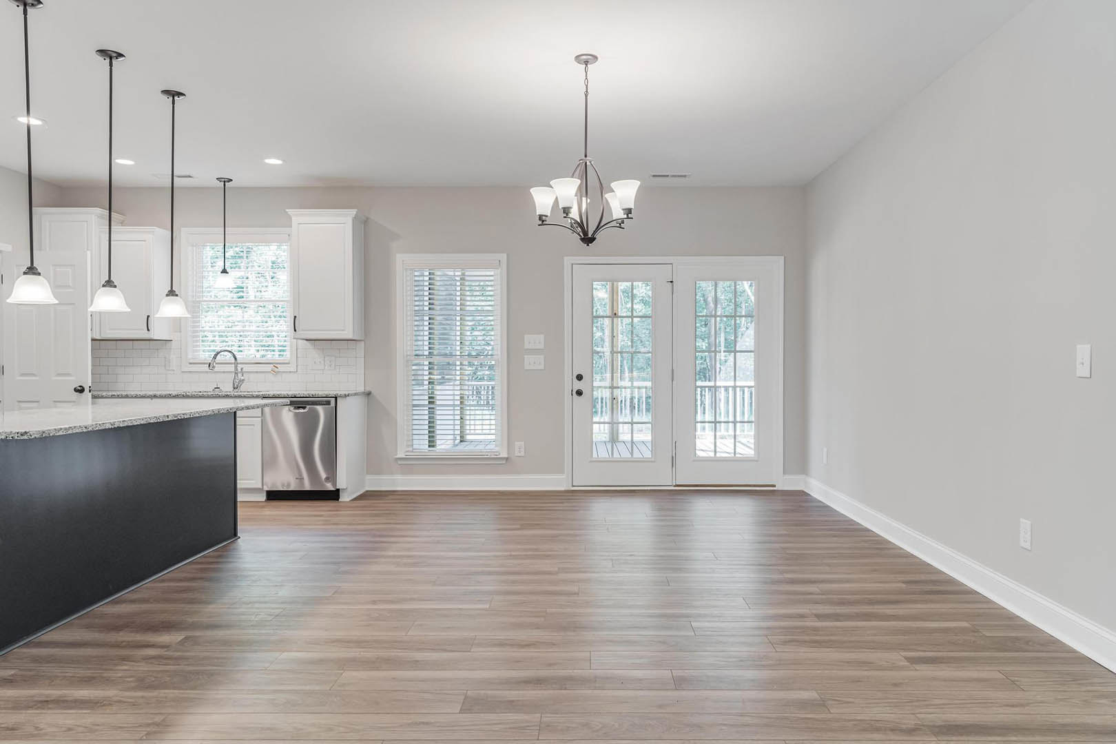 Open kitchen and dining area with hardwood floors, white double doors with glass panes, window with blinds, stainless steel dishwasher, and chandelier suspended from ceiling.
