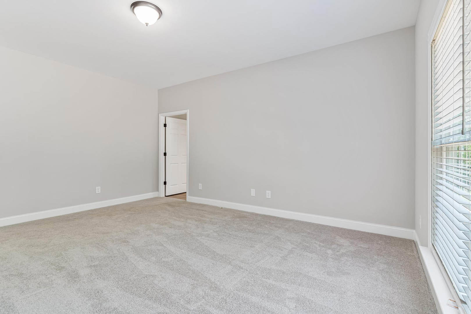 White paneled door with black handle, beige carpeted floor, white ceiling with flush mount light fixture, window with white trim, smooth plaster walls