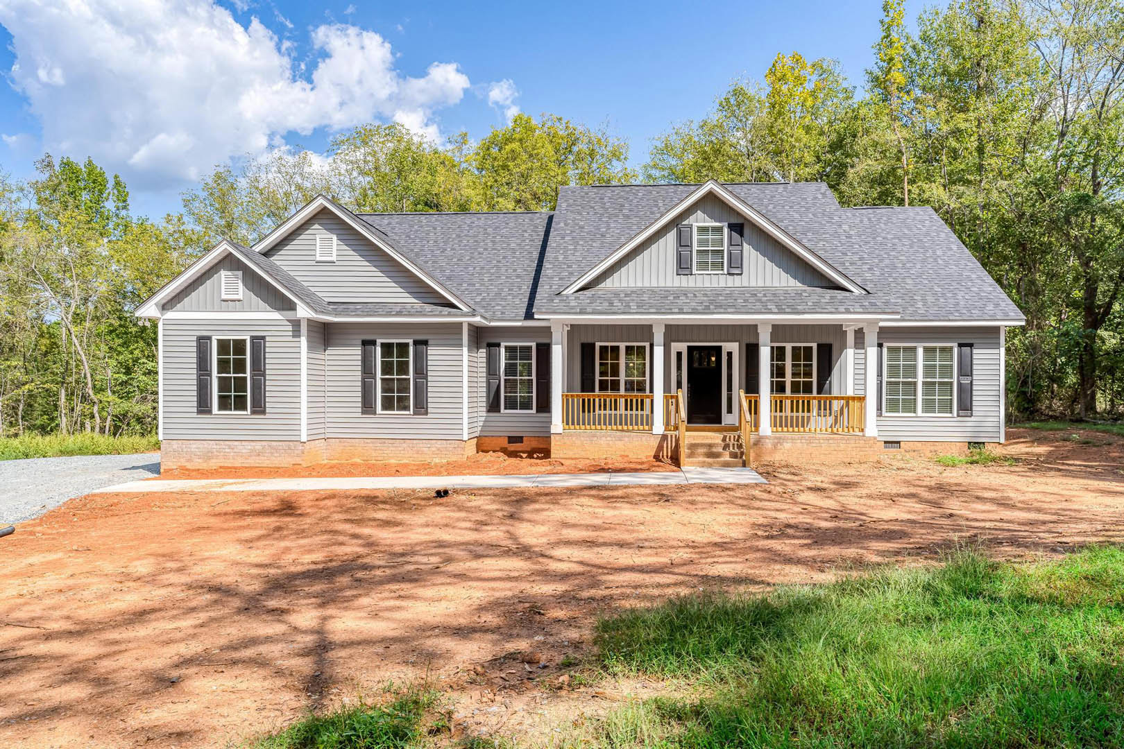 Two-story house with white siding, black front door, white-framed windows, covered front porch, dirt driveway, grassy yard, and mature trees surrounding the property