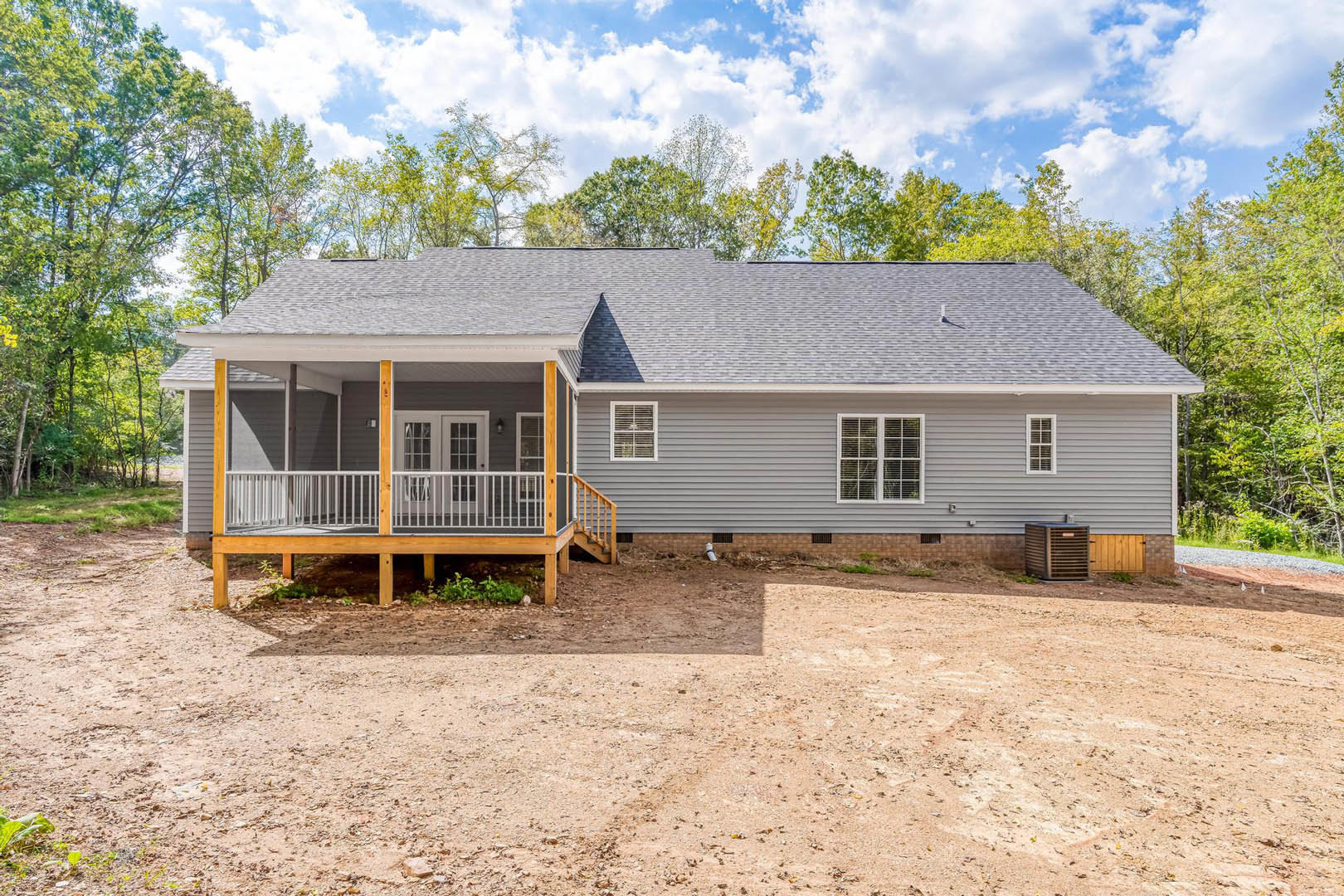 Two-story cottage with covered porch, white railing, stairs, dirt driveway, outdoor air conditioner unit, windows with blinds, surrounded by trees and cloudy sky.