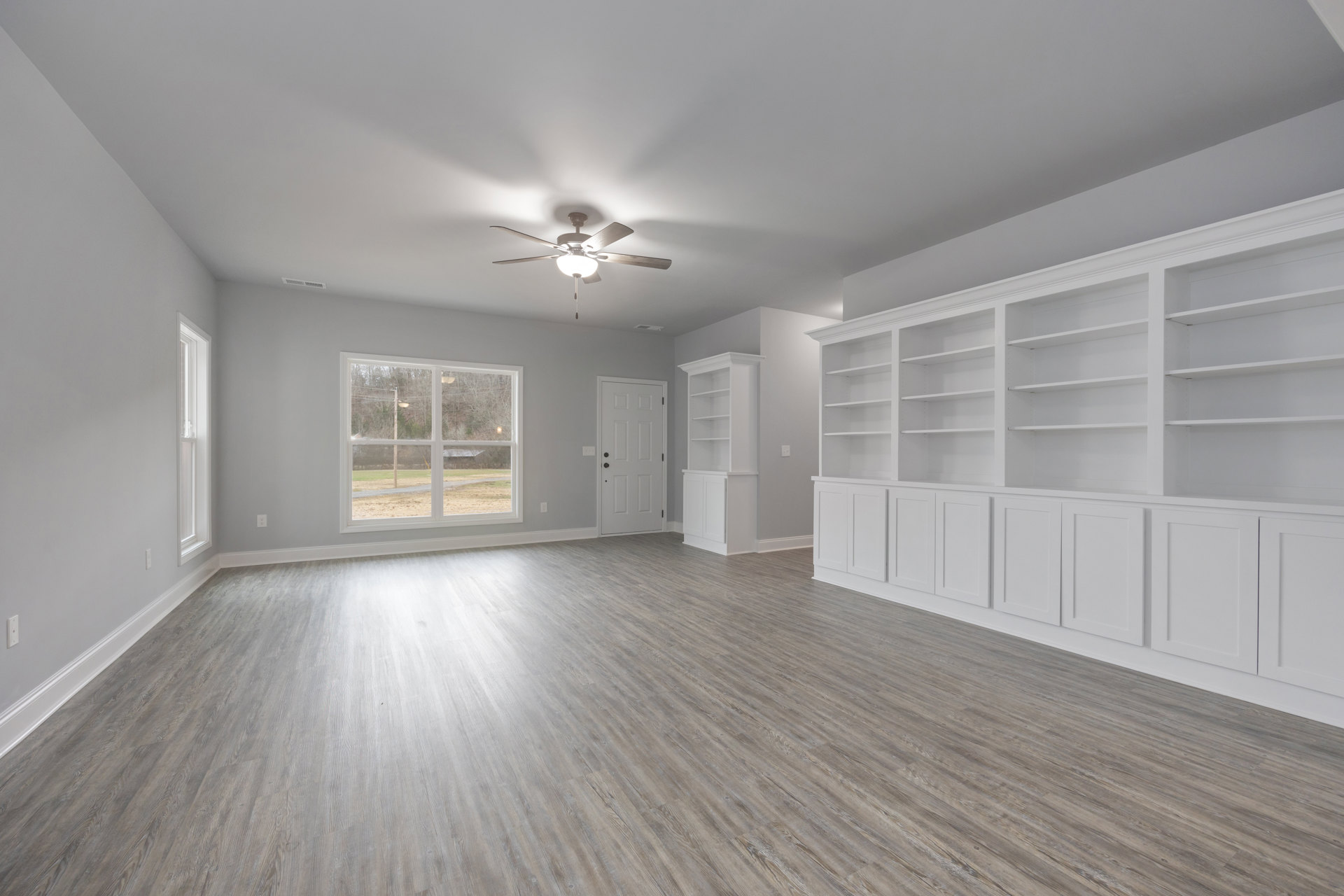 White built-in shelves against white walls, wood flooring, ceiling fan with light fixture, window overlooking field and trees, white door with black hardware