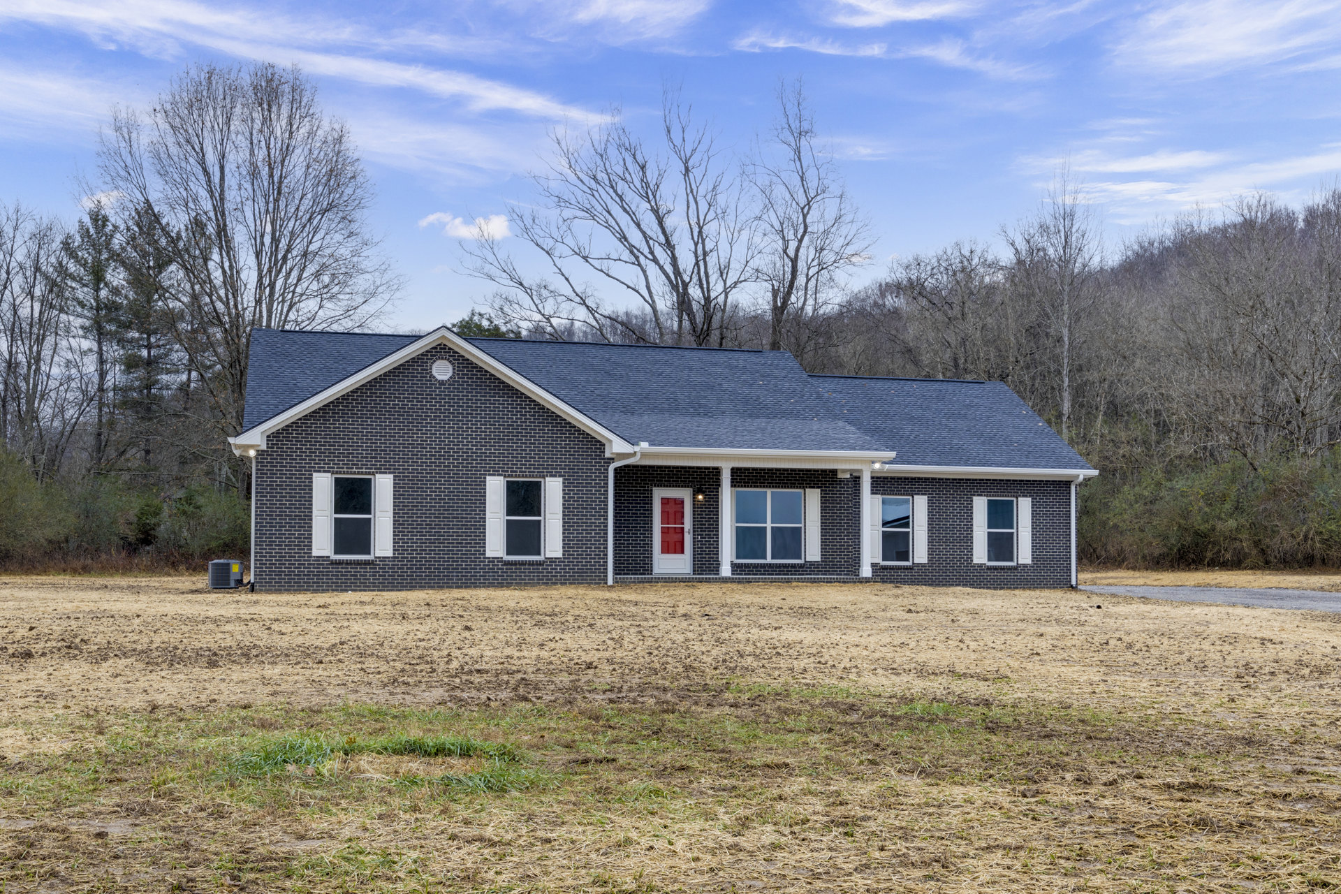 Brick house with white-trimmed windows and red door, surrounded by a large grassy yard, trees and dirt field in the background, cloudy sky overhead