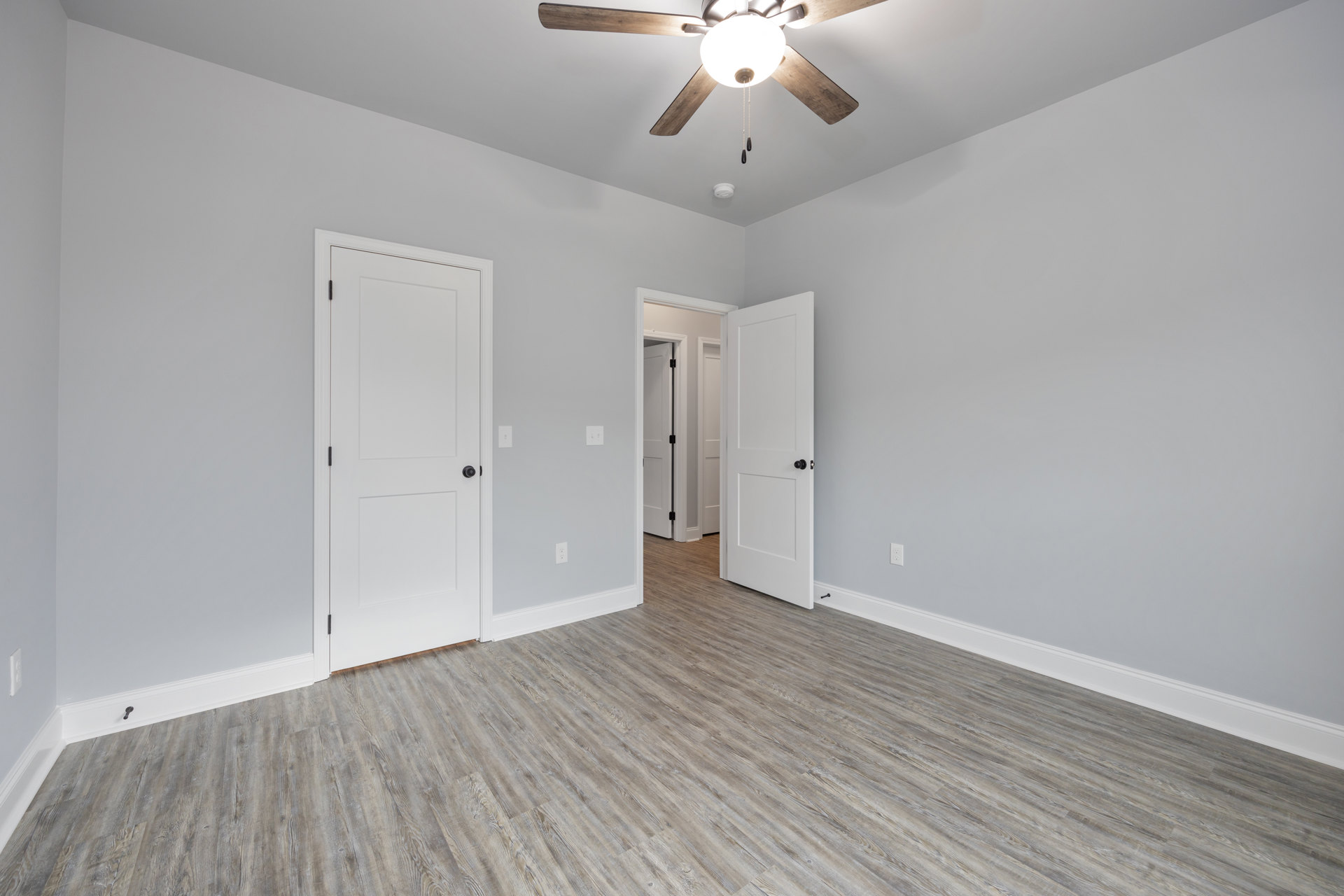 Wood floor and white walls in a room with two white doors featuring black hardware, ceiling fan with light fixture, crown molding, and plaster ceiling.