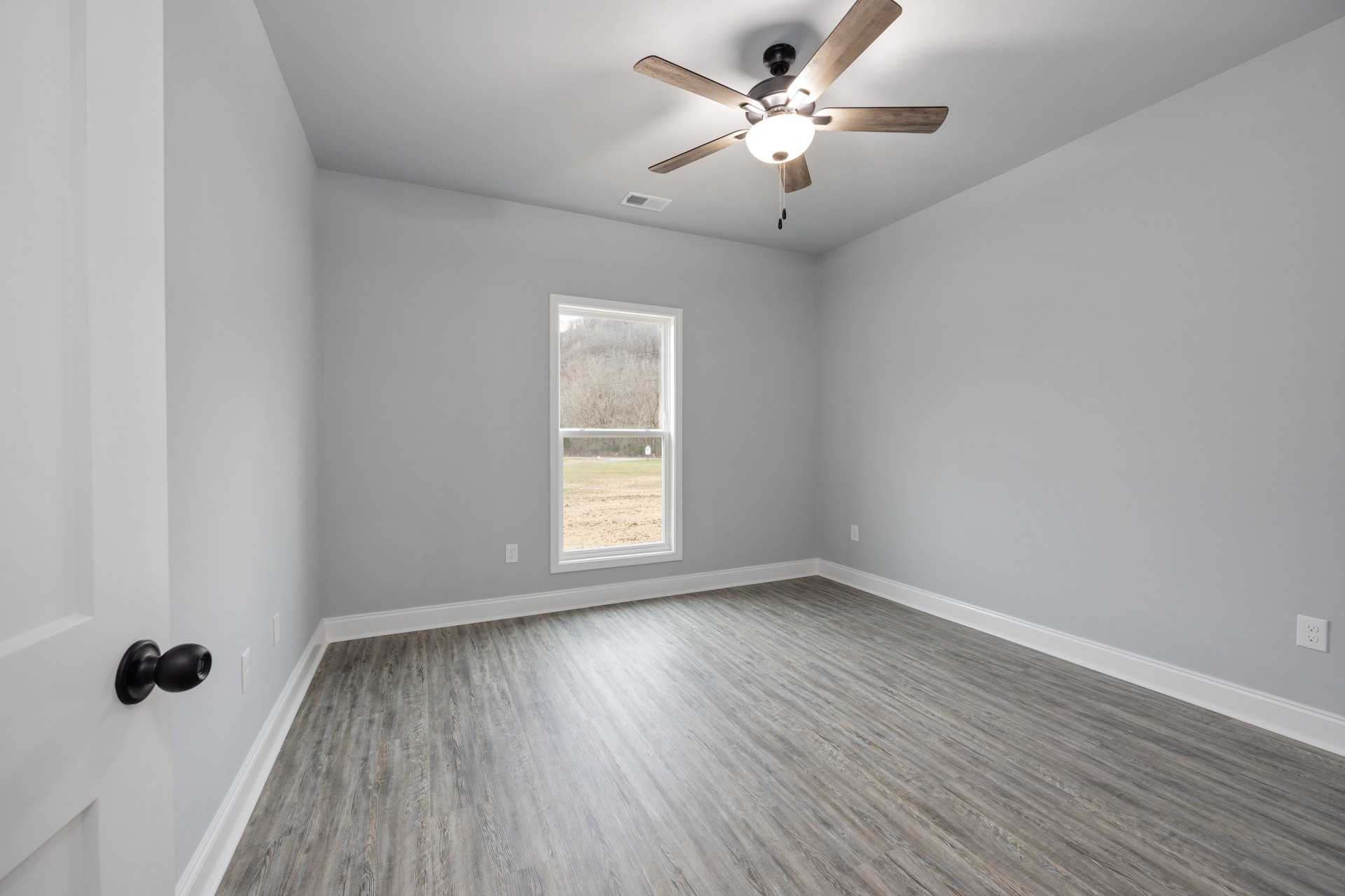 Wood-floored room featuring a ceiling fan with light fixture, large window overlooking trees and grass, plaster walls, and a black round door knob