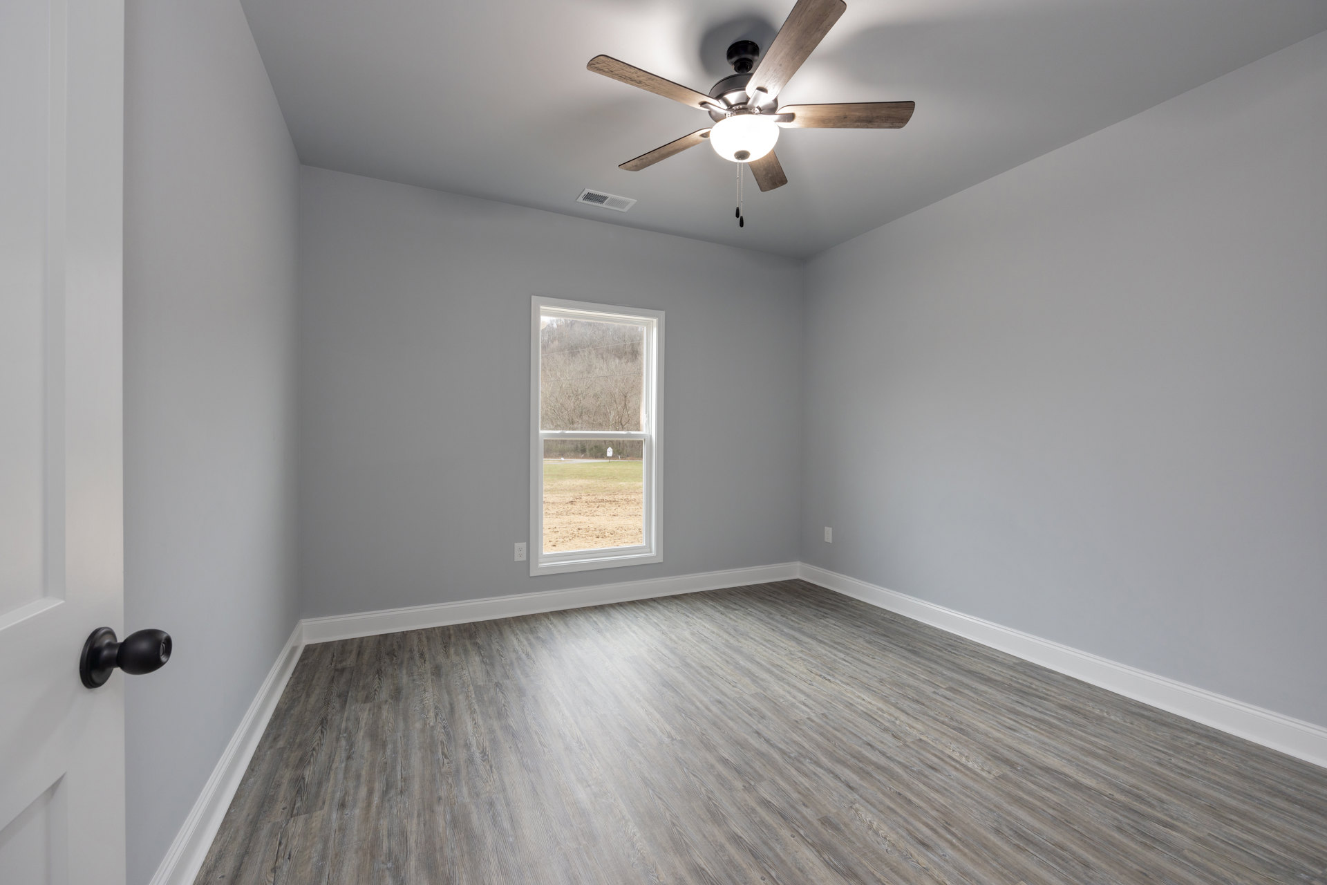 Ceiling fan with black light fixture on white ceiling, wood flooring, window overlooking trees and grass, white walls with molding, black round door knob