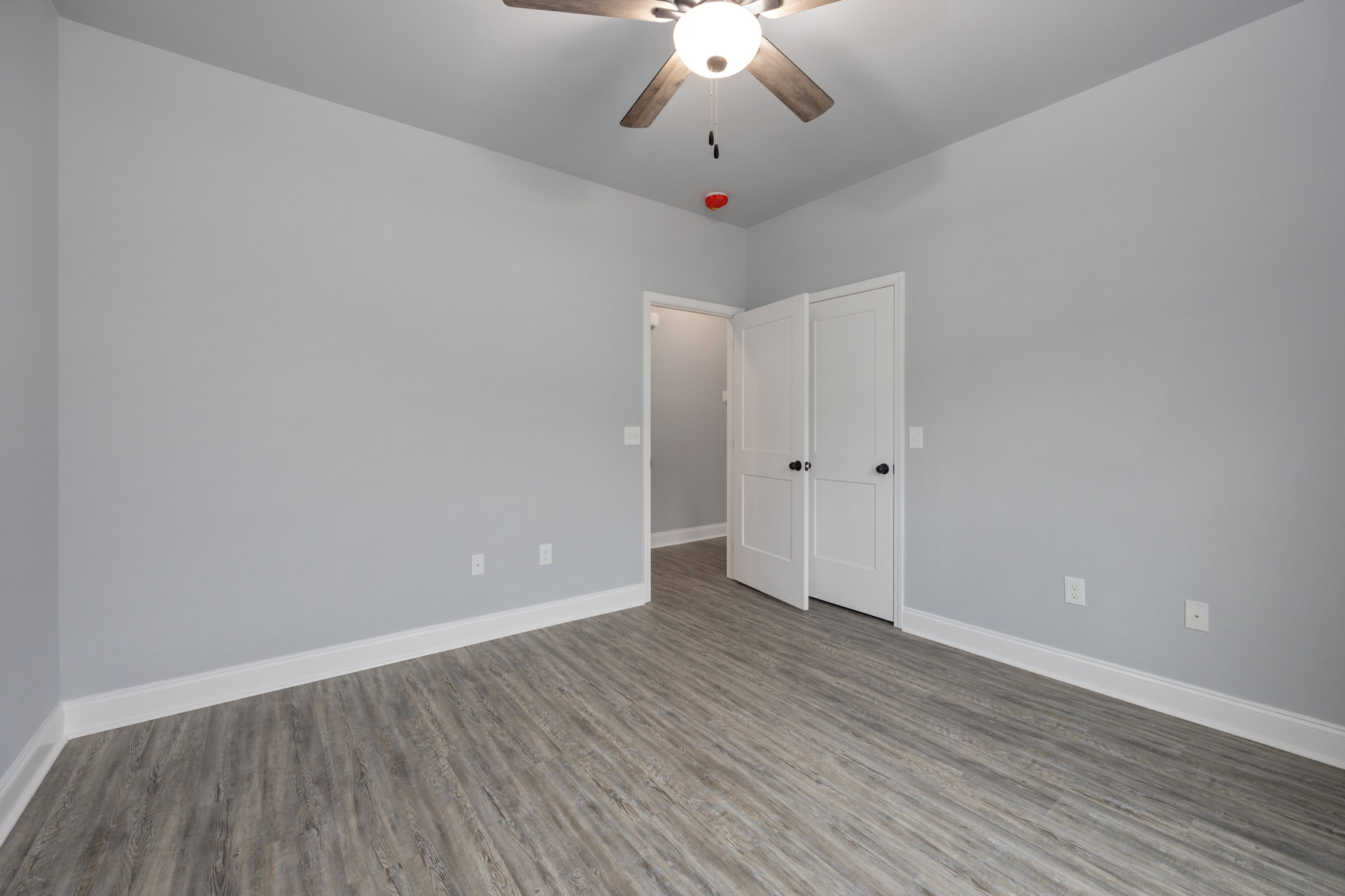 Ceiling fan with light fixture mounted on white plaster ceiling, wood laminate flooring, two white doors with black knobs, neutral walls.