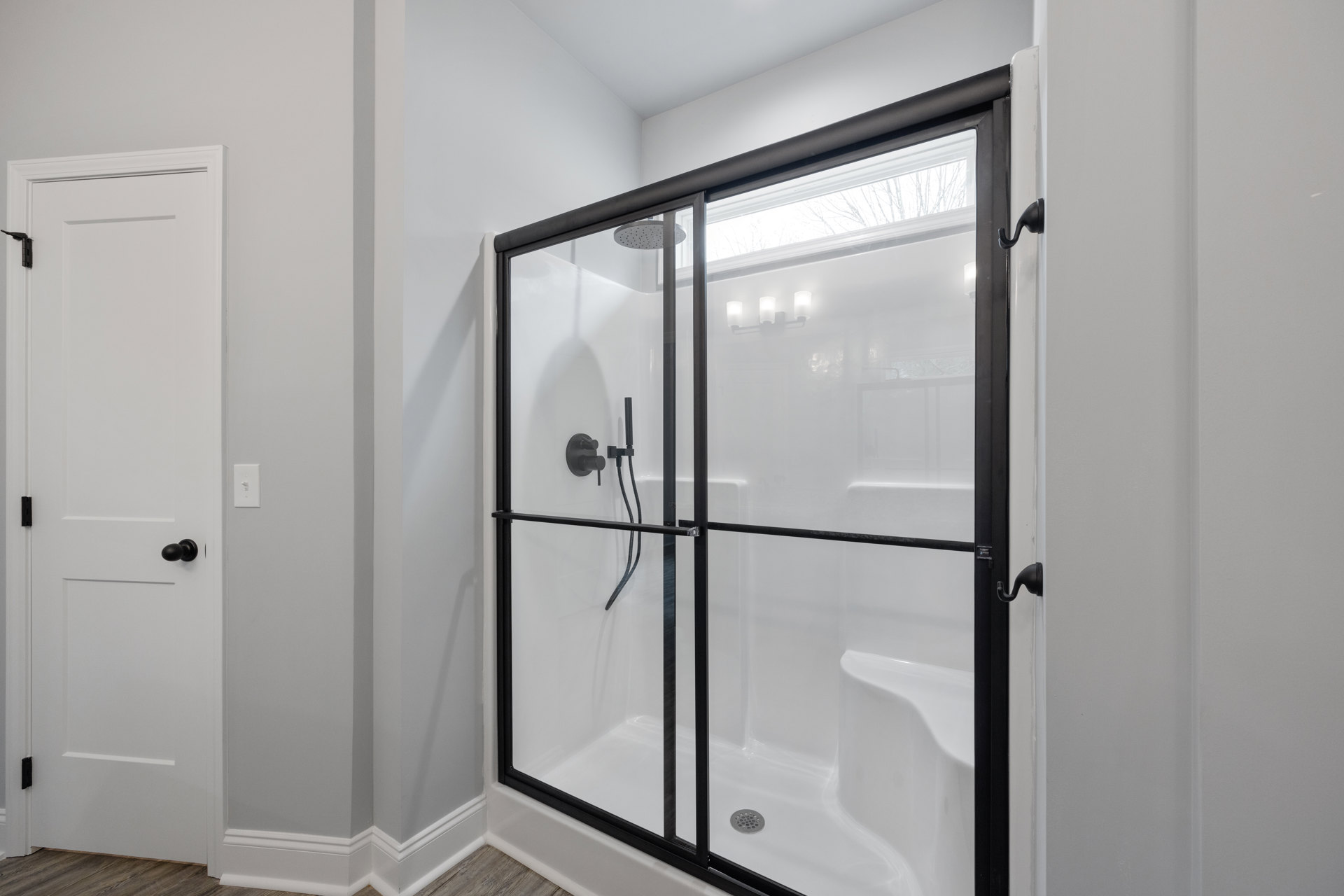 Walk-in shower with clear glass door, white tile walls, black hardware, adjacent white bathtub with black frame, white door featuring black knob and wall hook