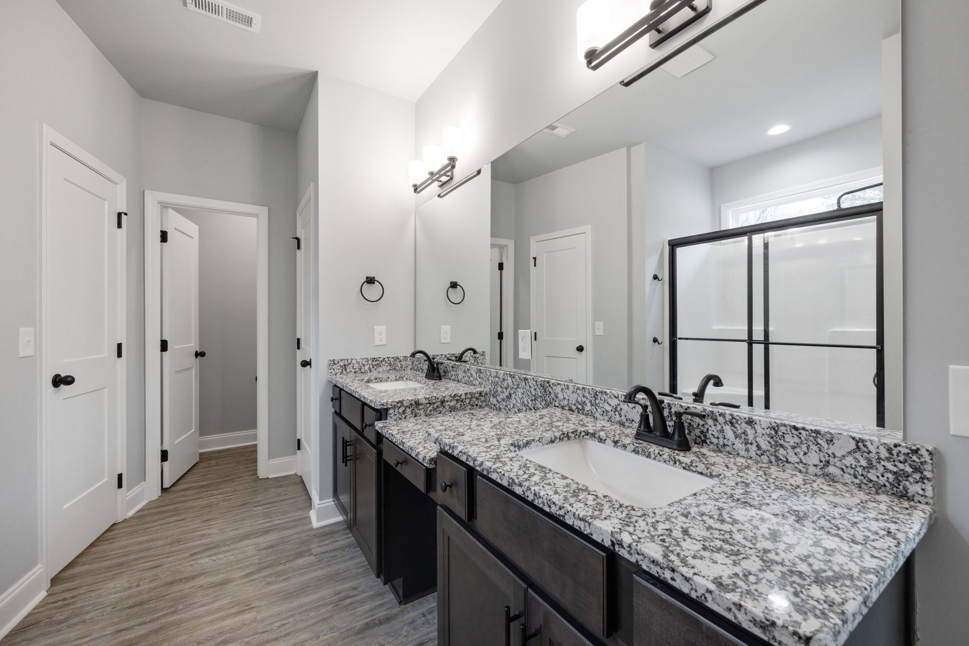 Bathroom with marble countertops, dual sinks featuring black faucets, white cabinetry, tile flooring, and a shower area near a window.