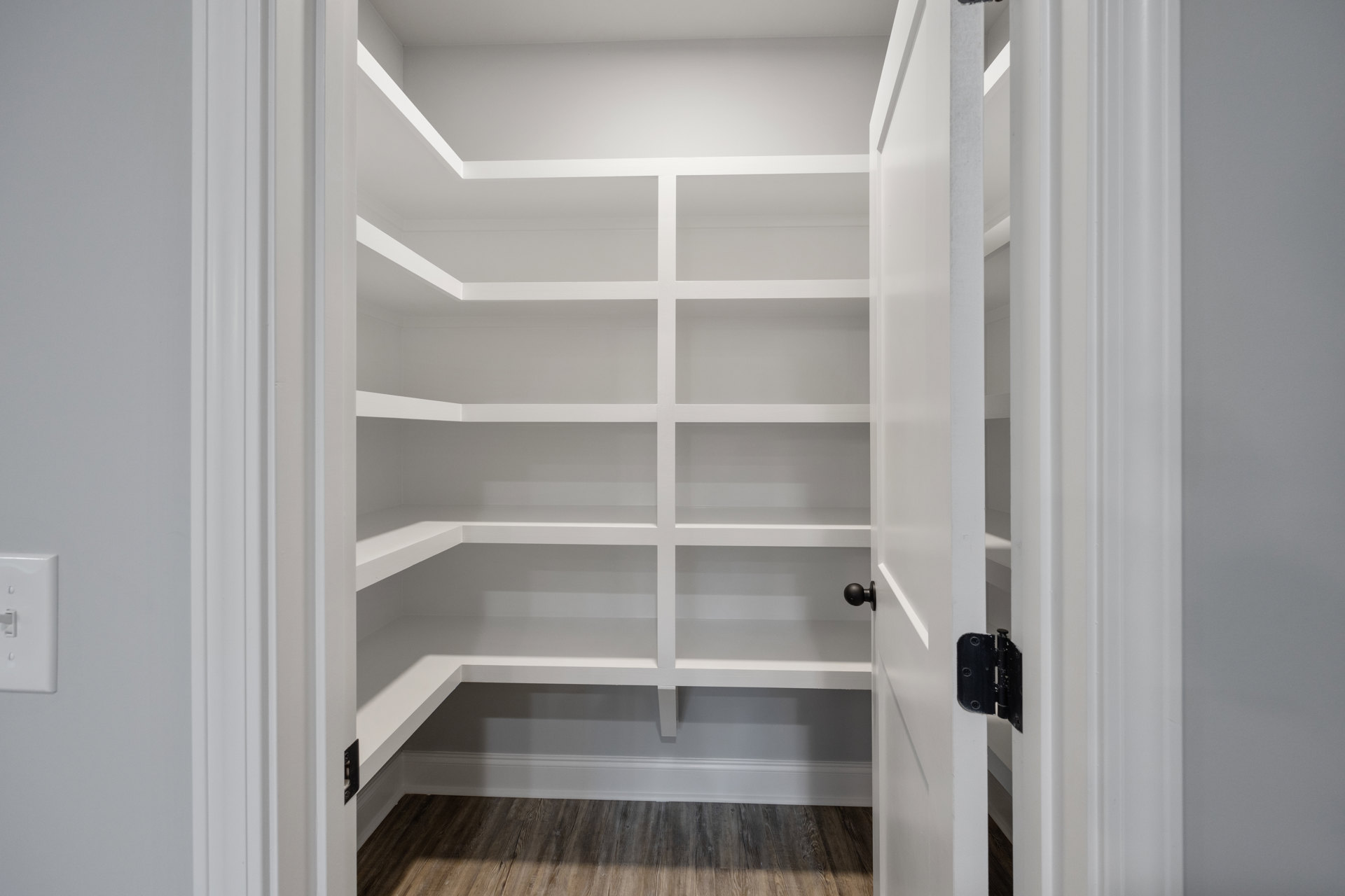 Open white door revealing closet with built-in white shelves, wood flooring, white baseboard trim, and black door hinge